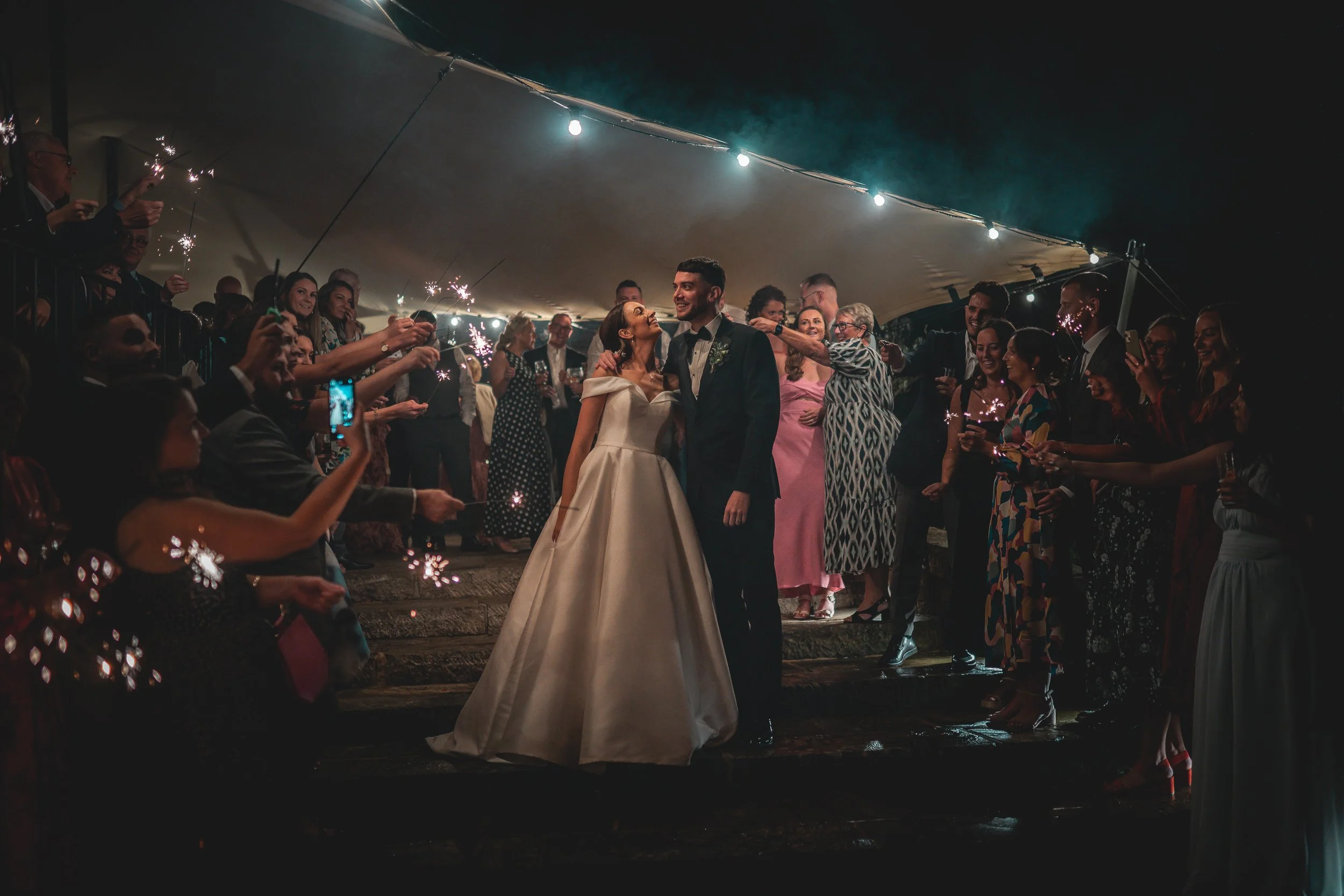 A bride and groom smiling at each other during their wedding celebration under a large tent with guests surrounding them holding sparklers.