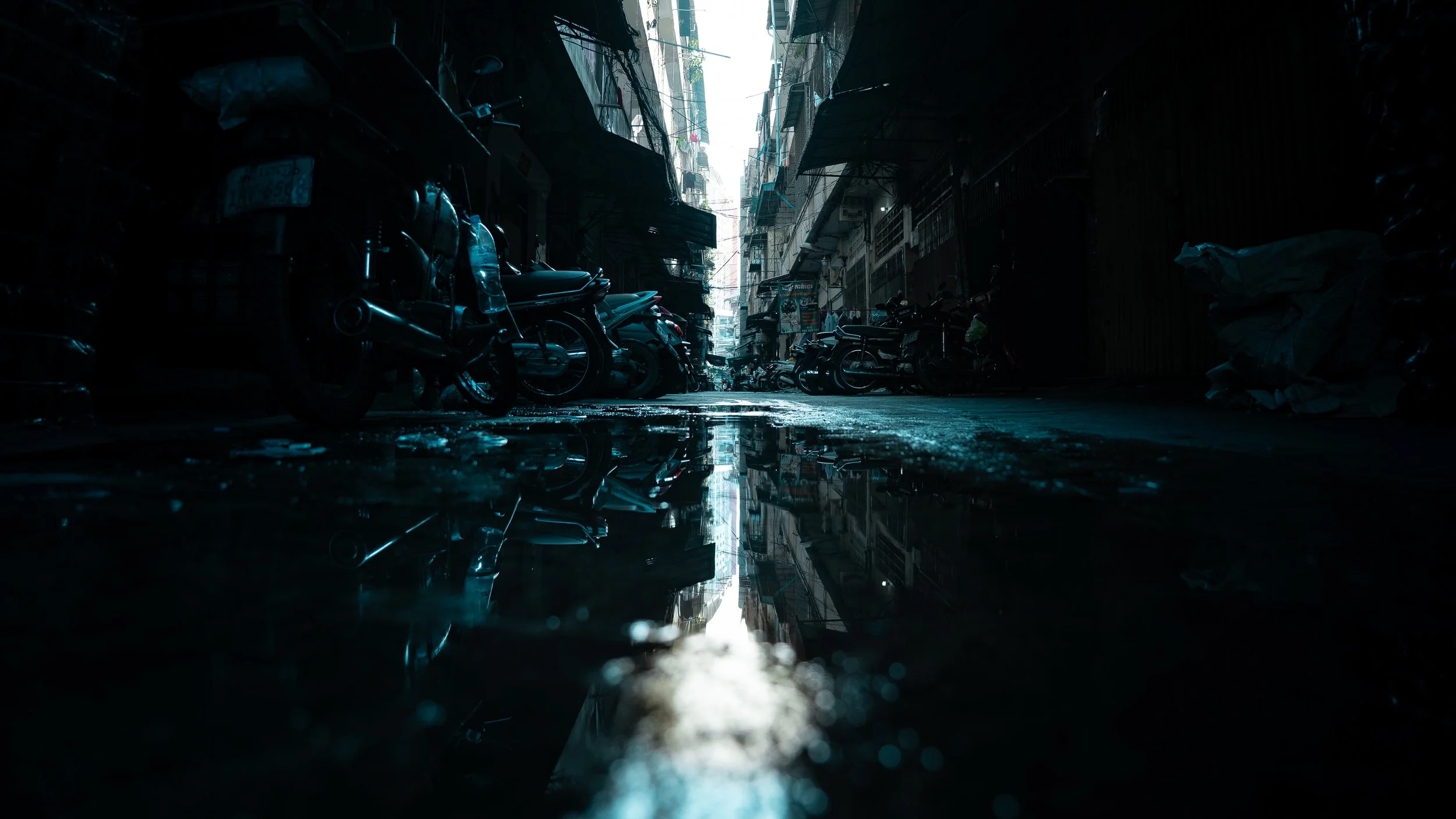 Dark alleyway with motorcycles parked along the sides, wet pavement reflecting the buildings above, and a glimpse of bright sky at the end of the alley.