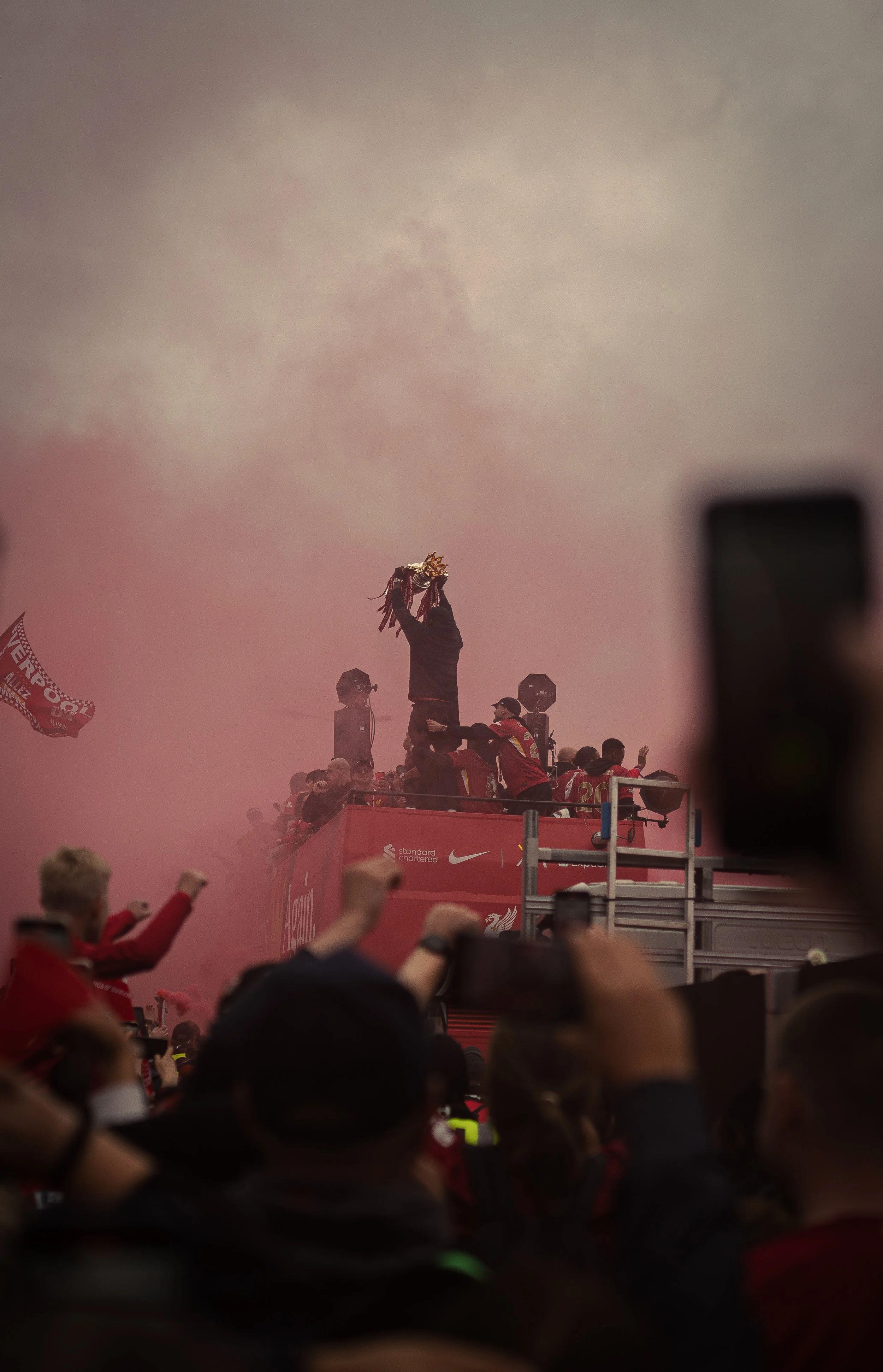 A celebration scene with a person holding a trophy high above their head on a red open-top bus surrounded by fans and smoke.