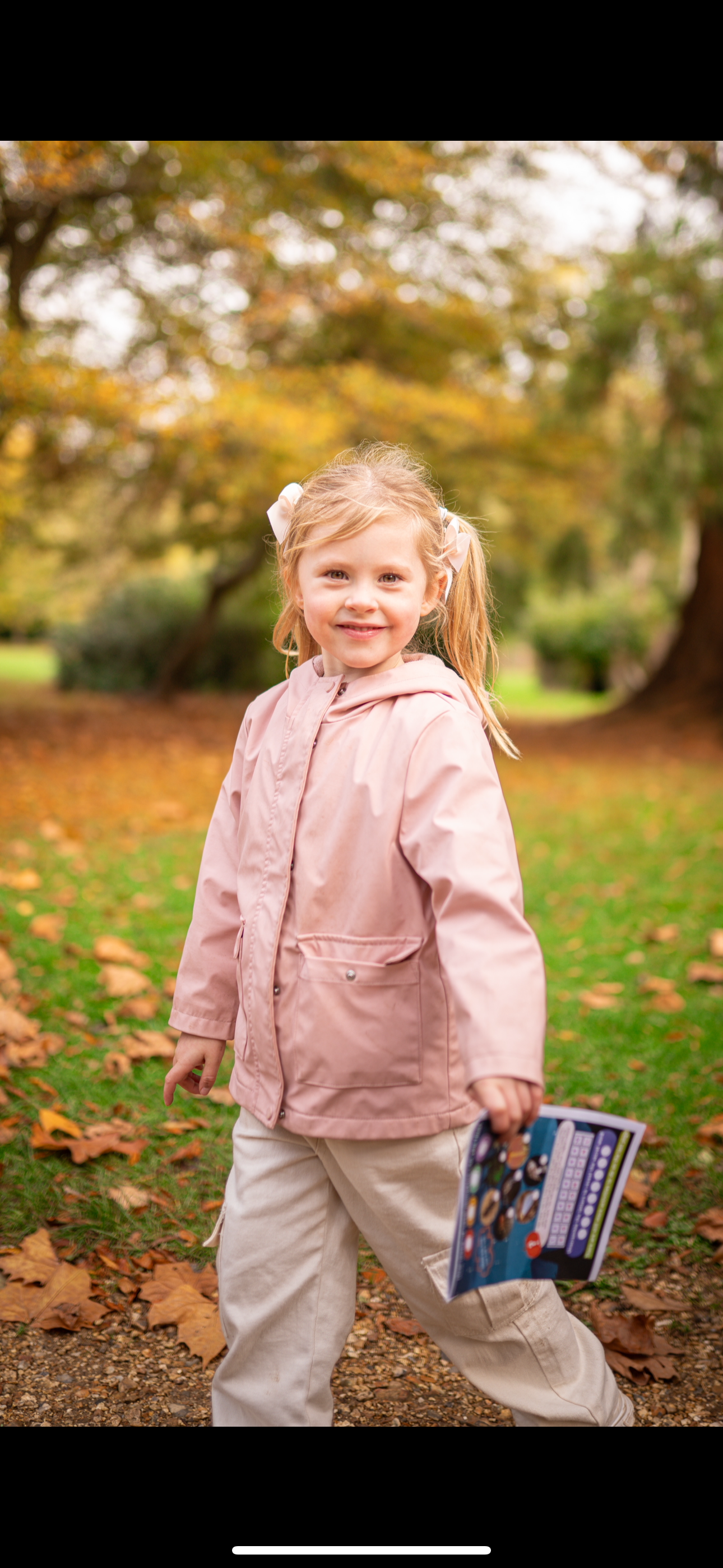 A young girl with blonde hair tied in pigtails with white bows, smiling, wearing a light pink jacket and beige pants, holding a tablet, standing outdoors among fall foliage.