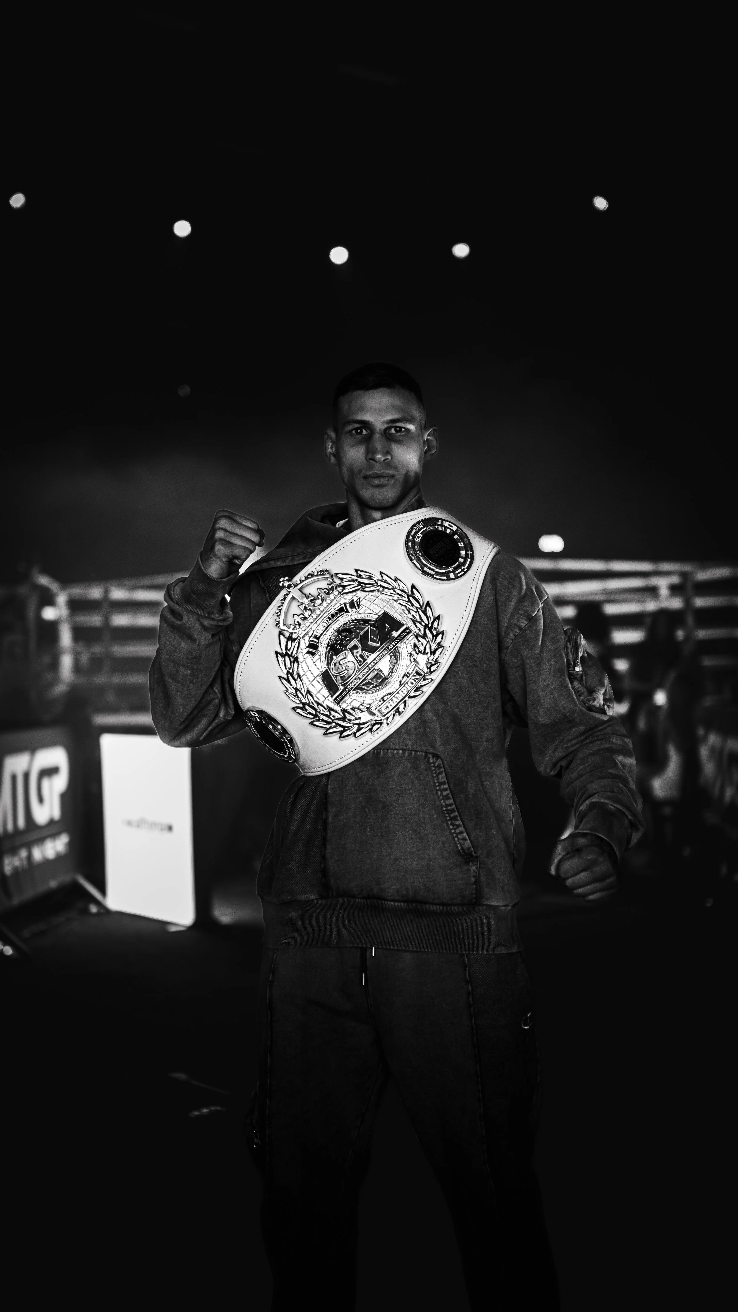 A young man dressed in casual clothes with a championship belt over his shoulder, standing in a dark, indoor setting with spotlights overhead, posing with his fist raised.