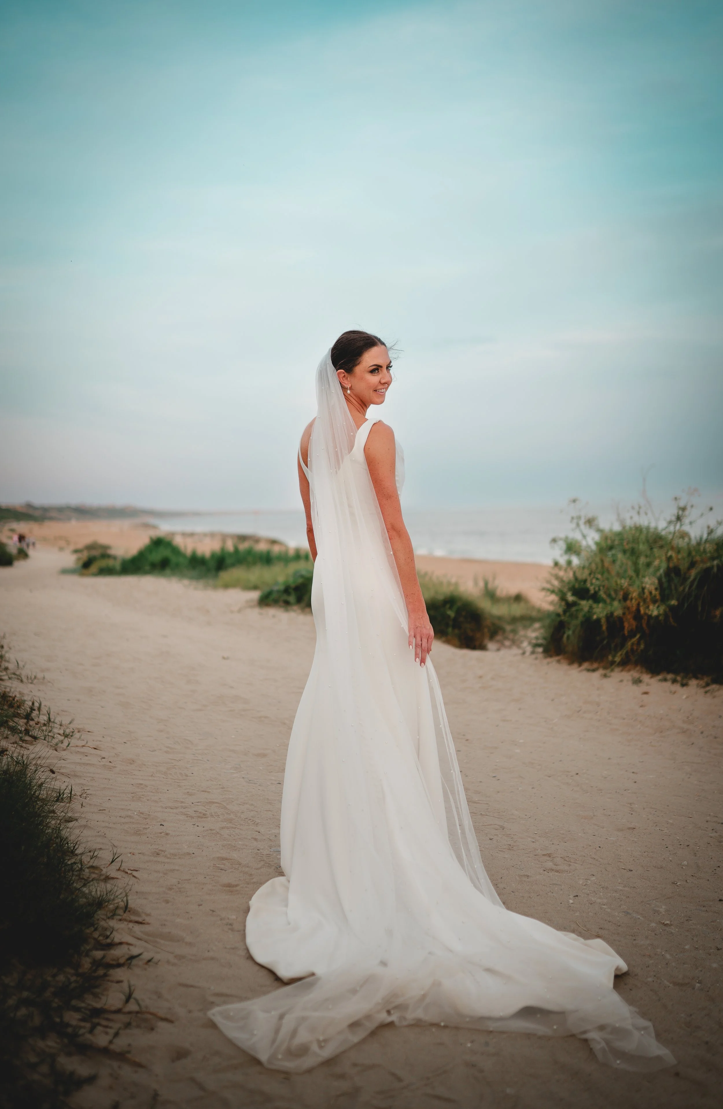 A bride in a white wedding dress and veil standing on a sandy beach, smiling and looking over her shoulder towards the ocean under a cloudy sky.