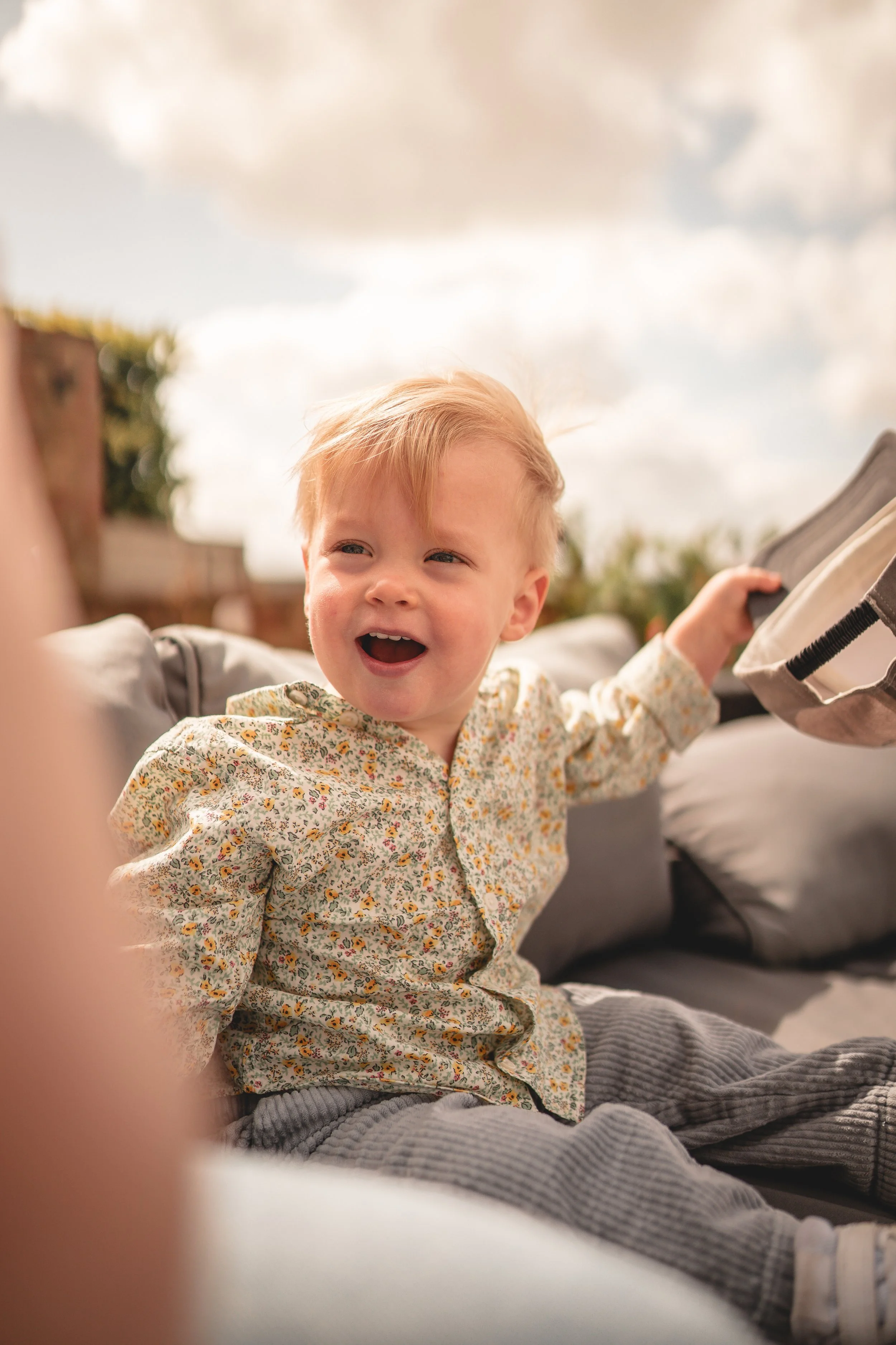 A young child with blonde hair sitting outdoors on a couch, smiling and holding a beige bag, with a cloudy sky and some plants in the background.