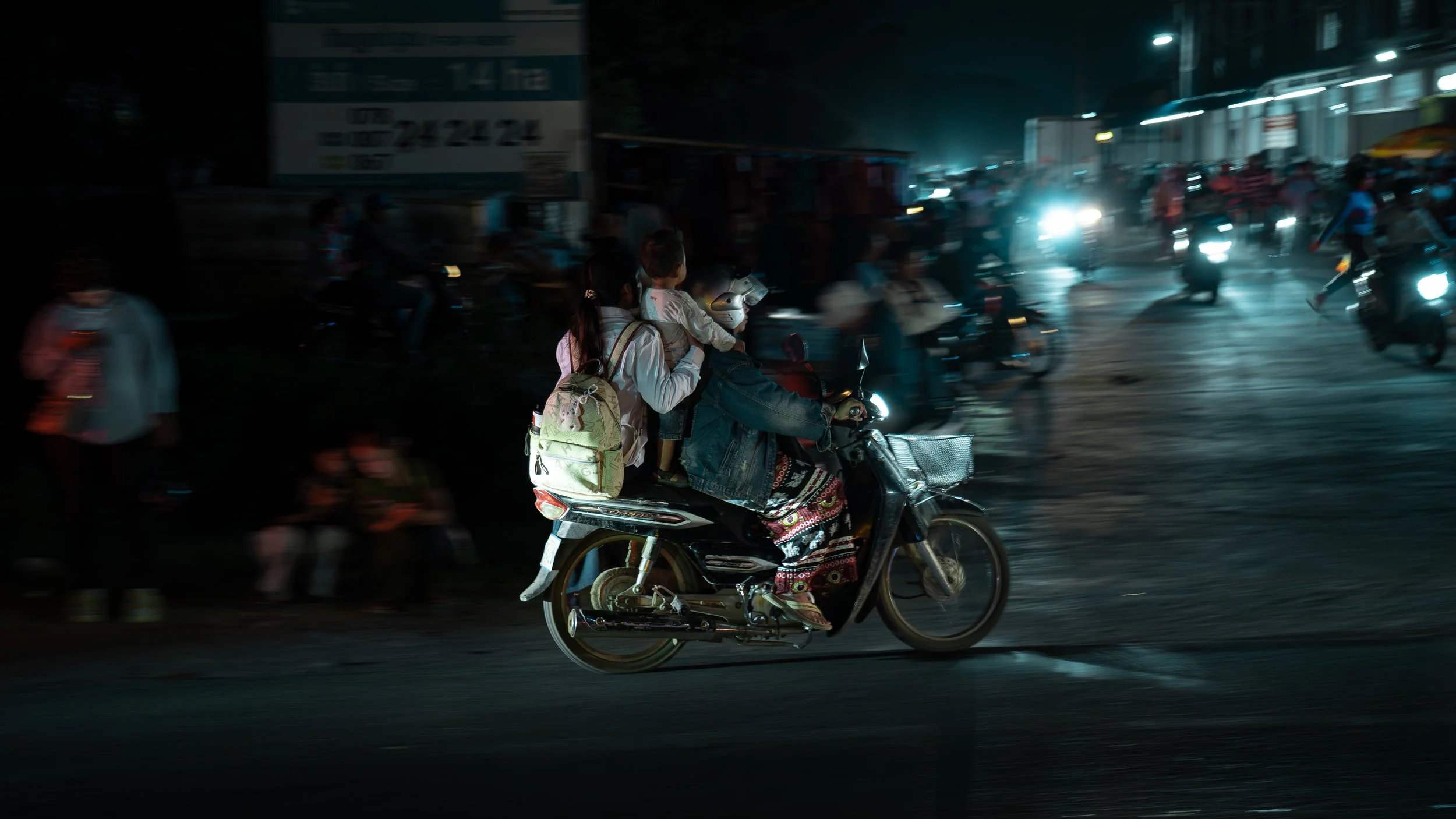A person riding a motorcycle at night with two children on board, one holding onto the rider and the other sitting behind, on a busy street with blurred vehicles and lights in the background.