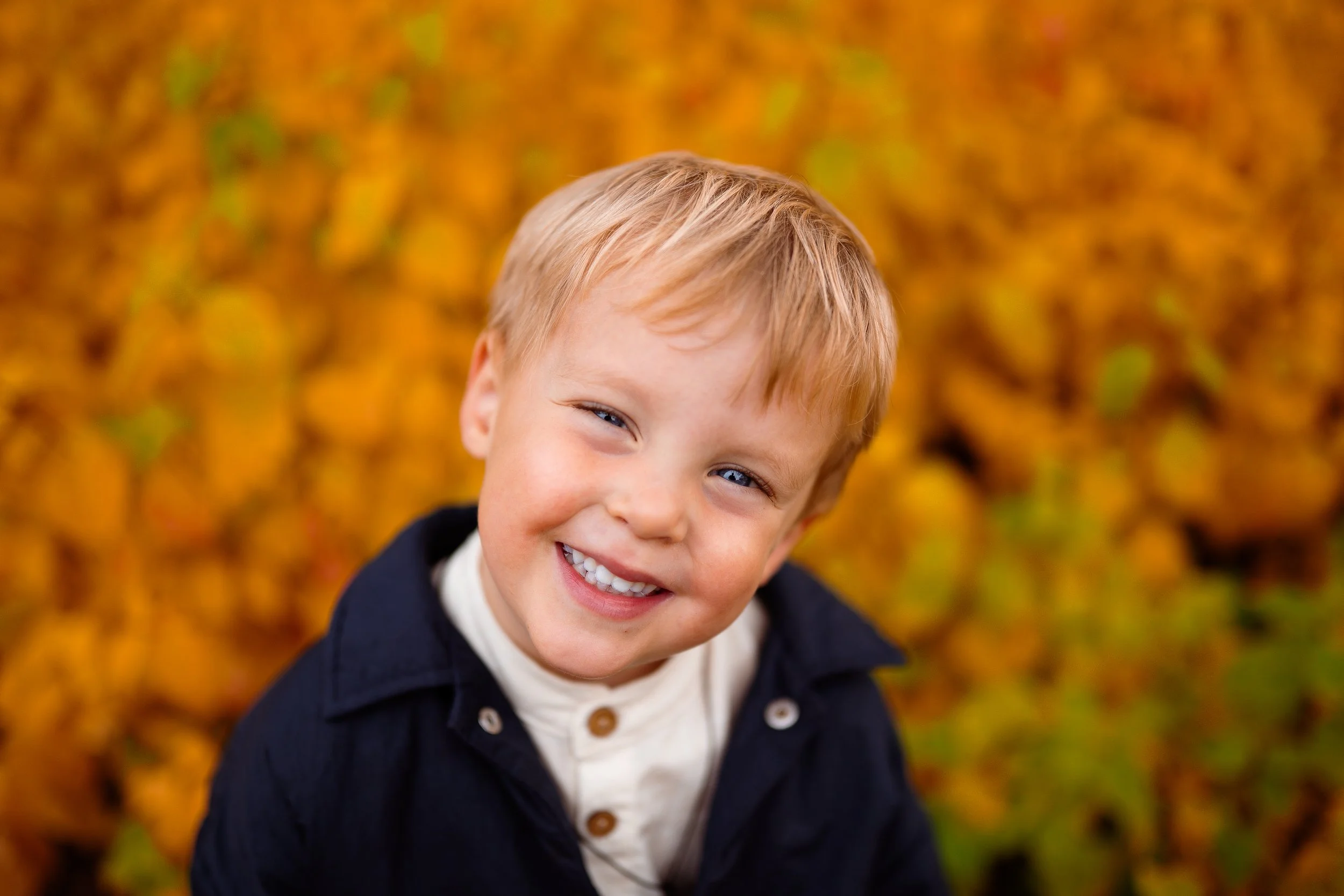 A young boy with blonde hair and blue eyes smiling outdoors in fall, with orange and yellow leaves in the background.