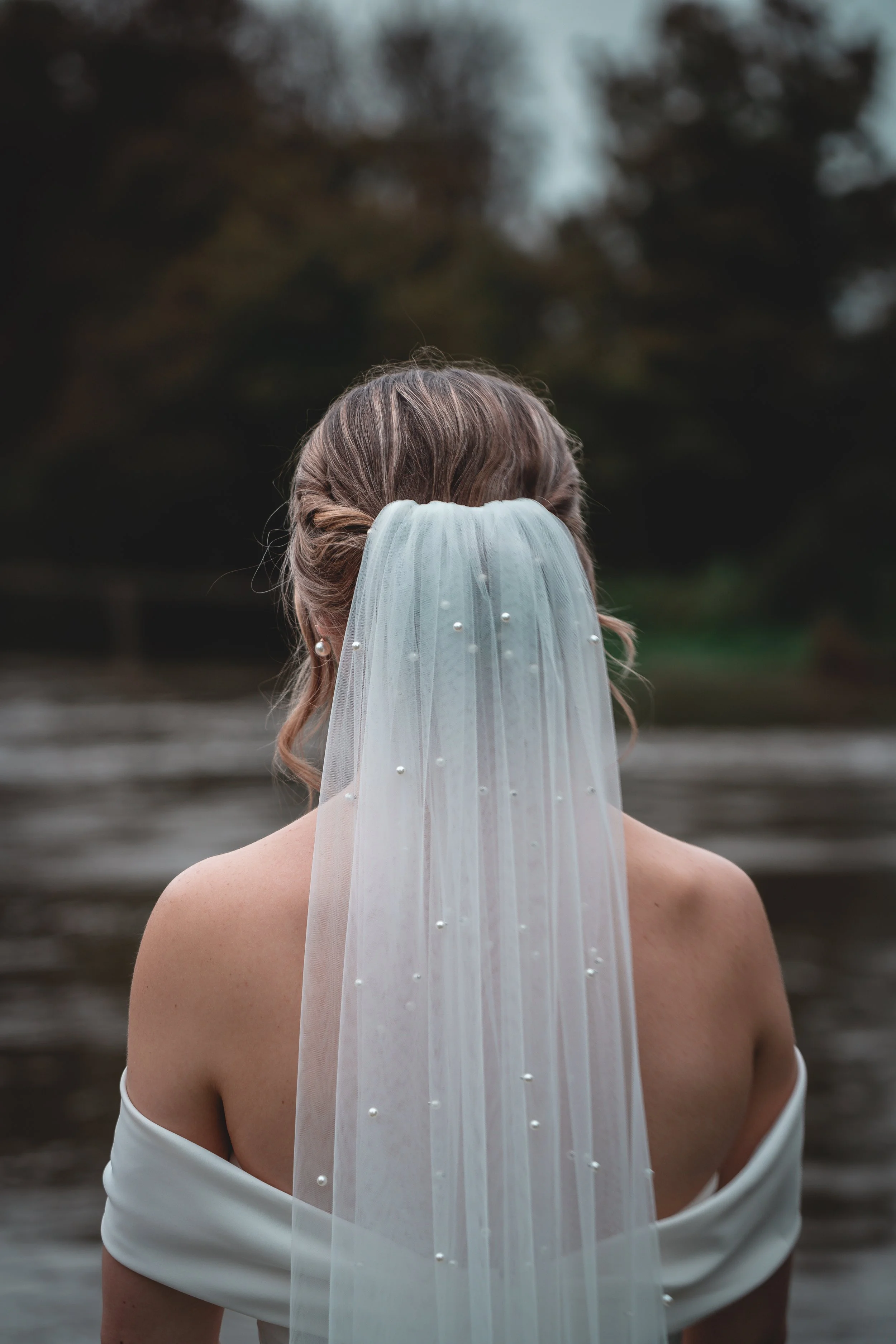 Back view of a bride with a veil and pearl details, standing outdoors near water.