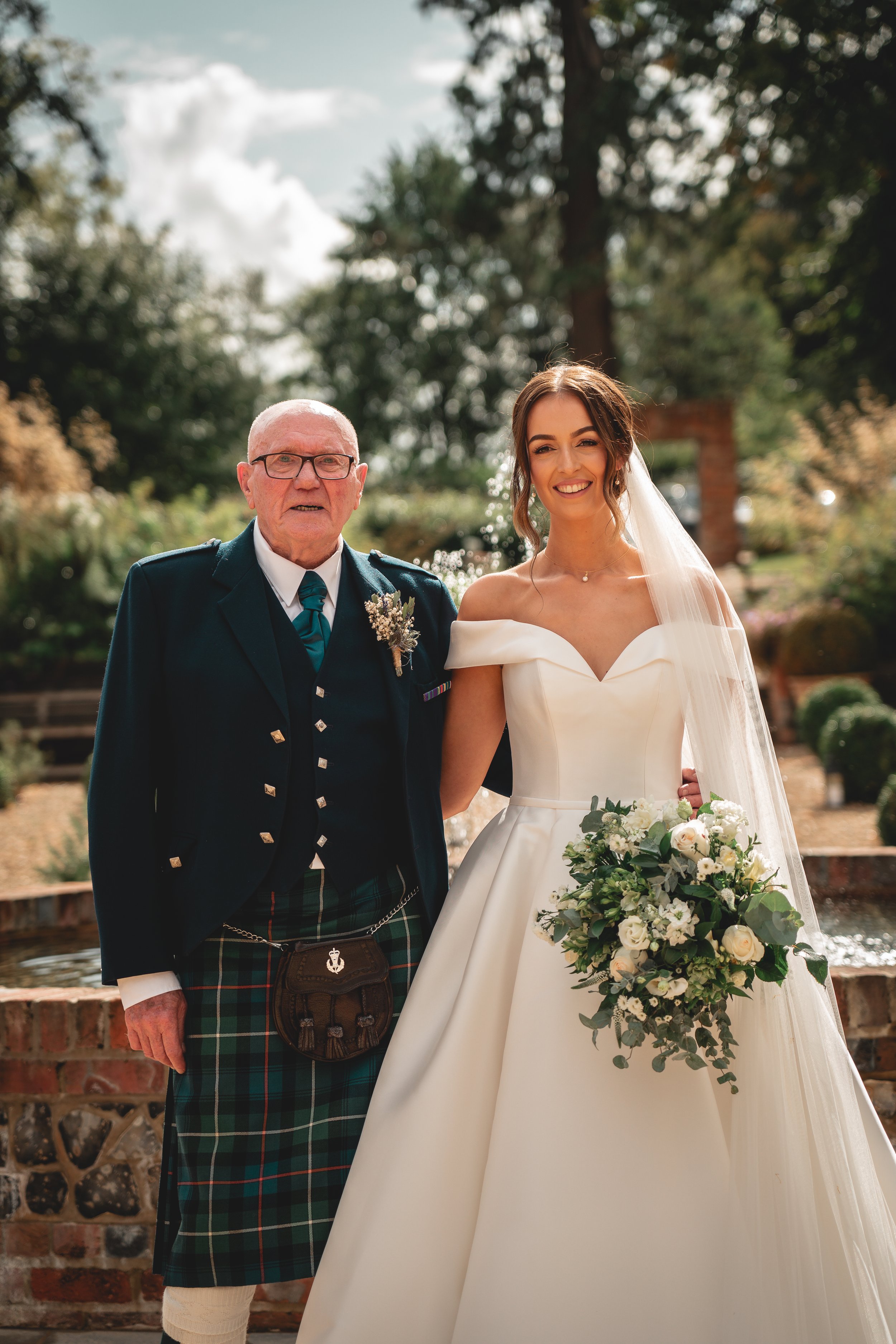 A bride in a strapless white wedding gown holding a bouquet of white and green flowers with an older man in a dark jacket, kilt, and glasses standing outdoors in a lush, sunny garden.