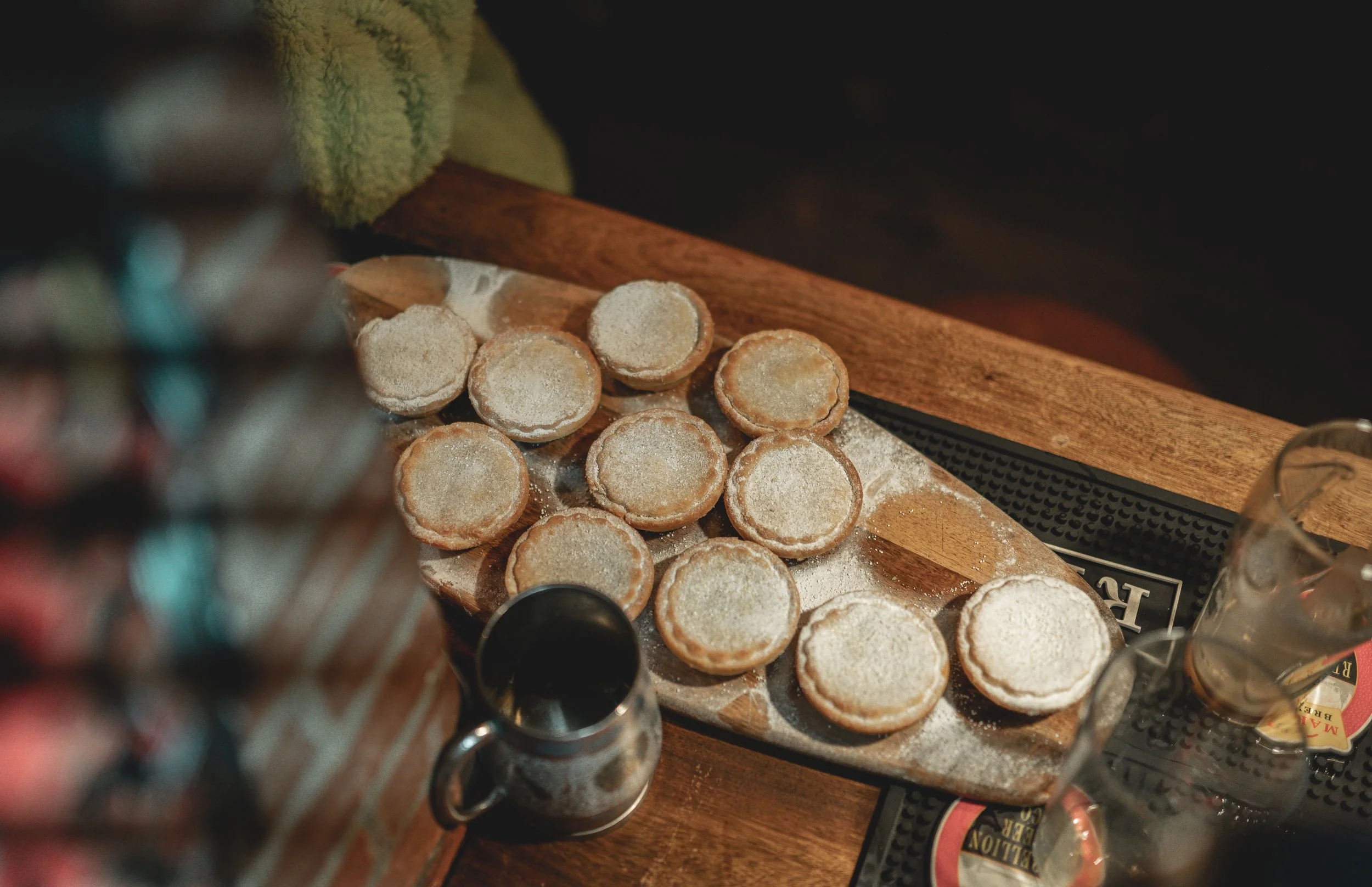 A wooden serving board with small round cookies dusted with powdered sugar on a table in a cozy setting, with a metal mug and glasses nearby.