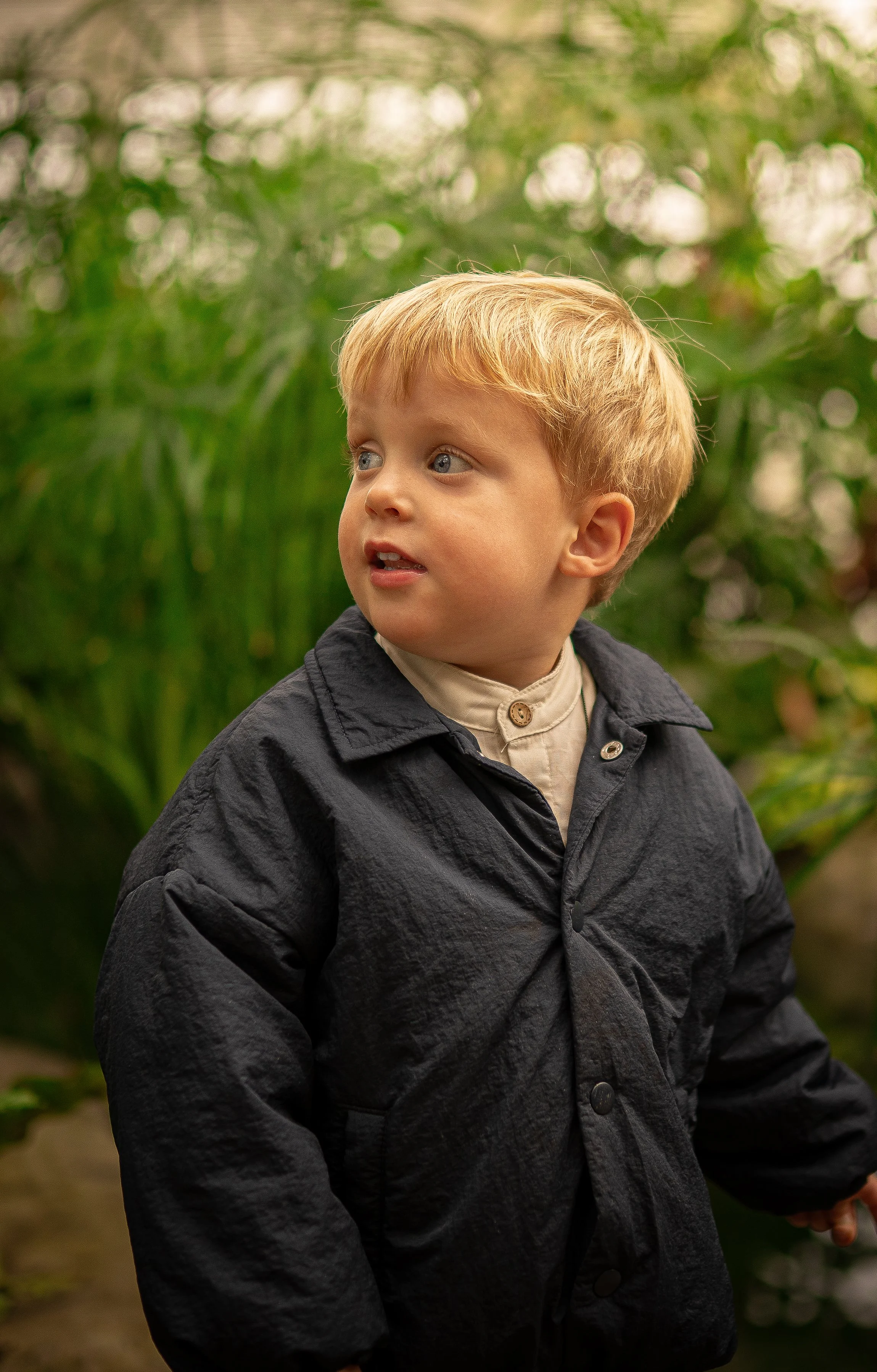 A young boy with blonde hair and blue eyes, wearing a black jacket and a beige shirt, standing outdoors with greenery in the background.