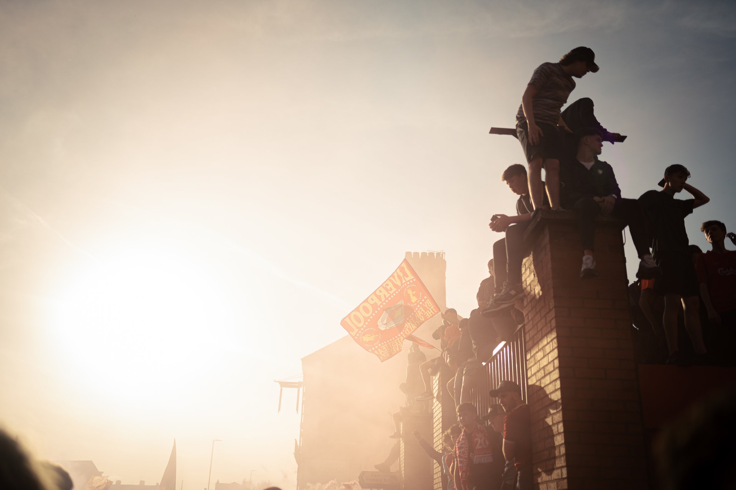 People gathered on a brick wall under bright sunlight, some sitting and some standing, with one person holding a flag. The scene appears to be outdoors during sunset or sunrise.