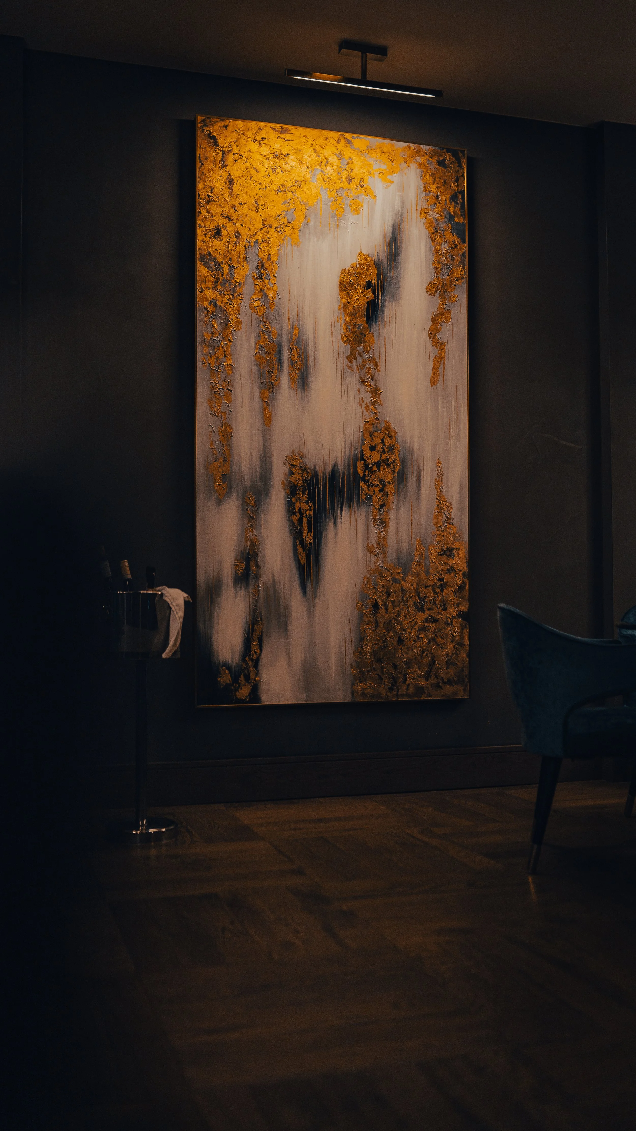 Abstract artwork with gold and black accents on a large vertical canvas, illuminated by a ceiling light, in a modern interior with dark walls and hardwood flooring.