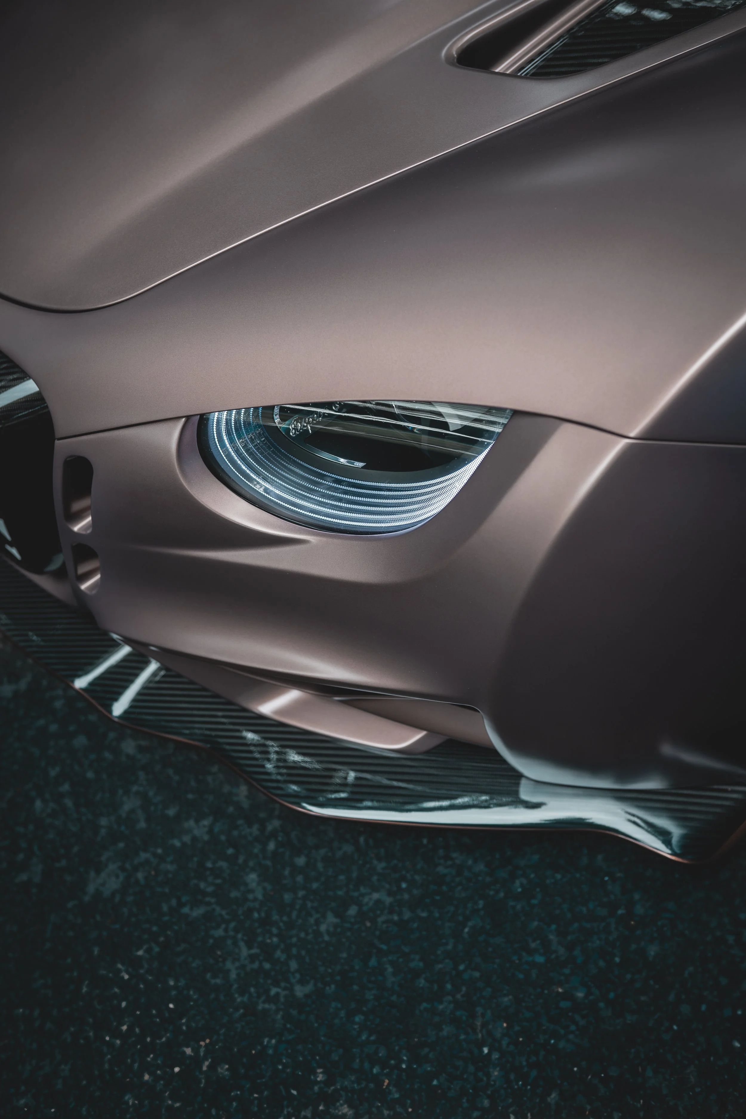 Close-up of a sleek, modern sports car's front, highlighting the headlight and aerodynamic design features on a dark asphalt surface.