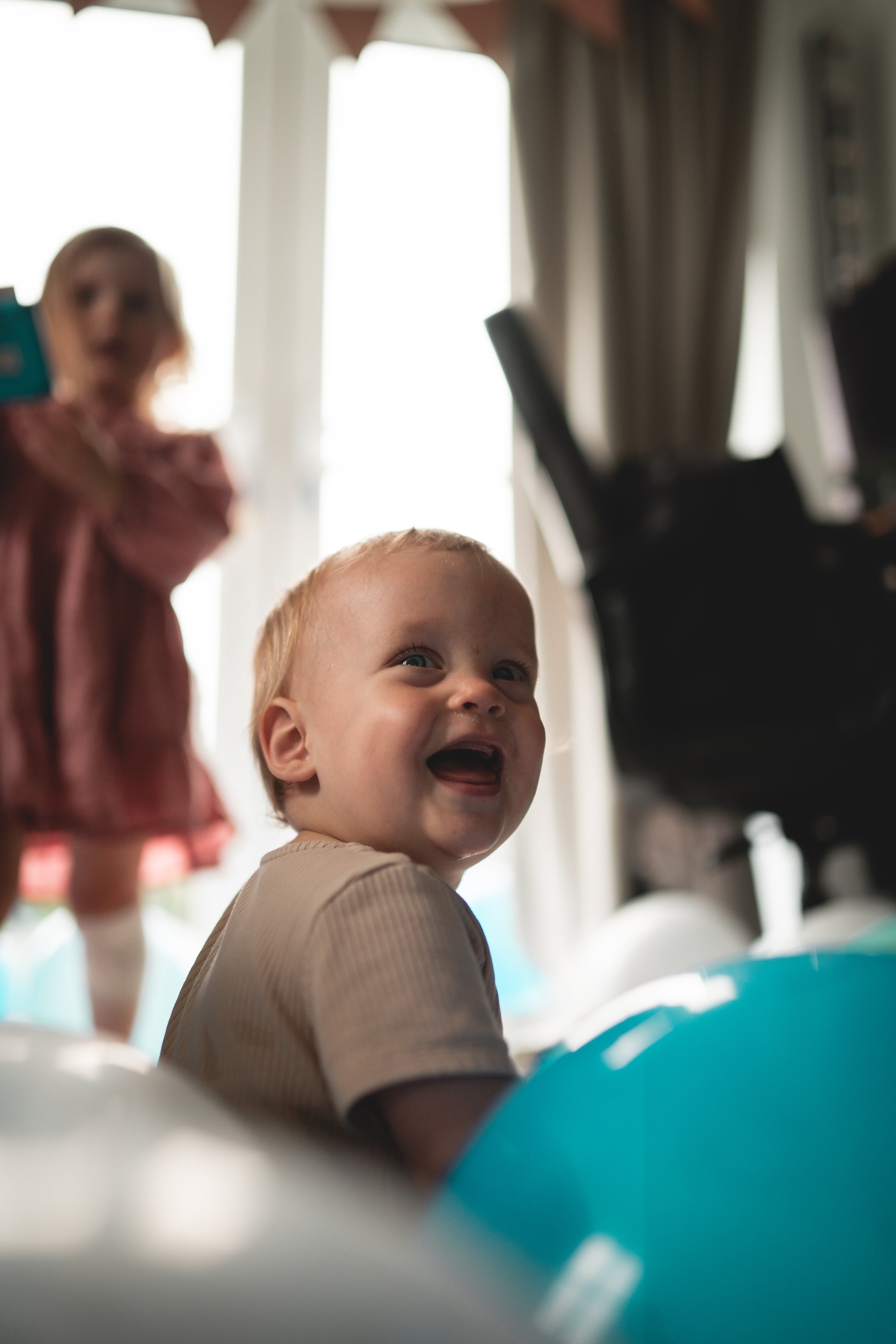 Smiling young boy with light hair sitting among balloons in a brightly lit room, with a girl in the background holding a phone.