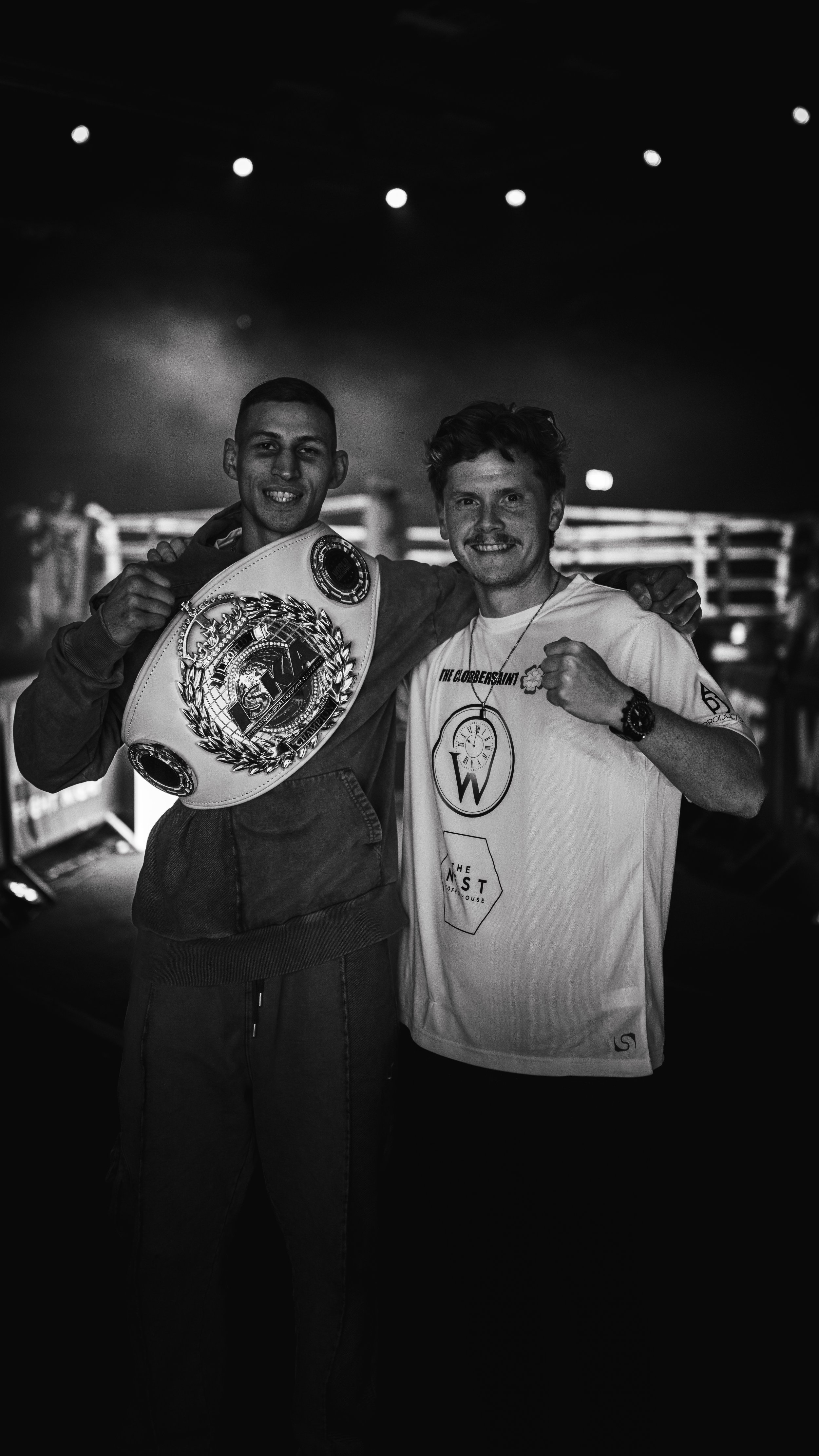 Two men smiling, one holding a championship boxing belt, standing together in a dimly lit venue.