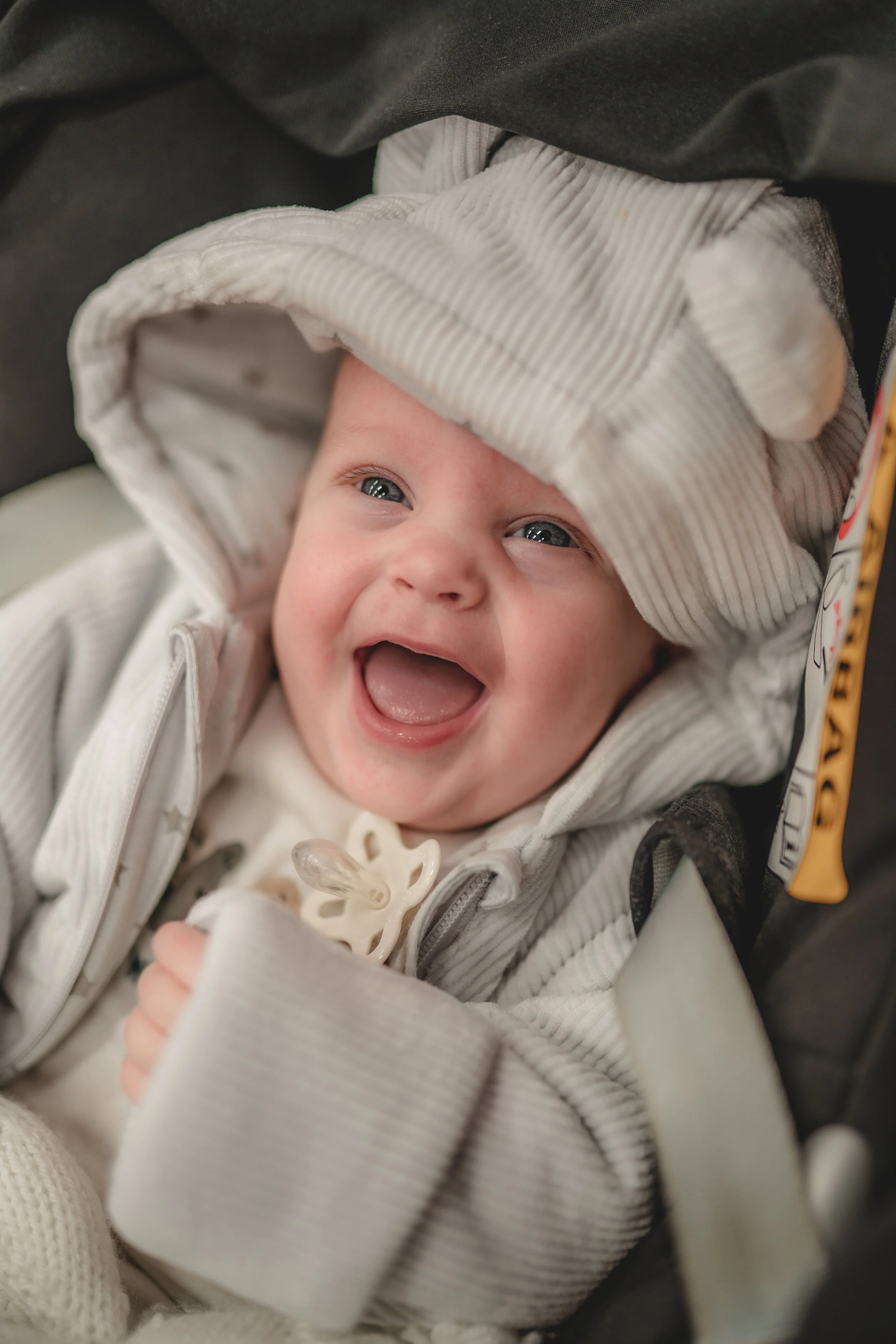 A smiling baby with blue eyes lying in a car seat, wearing a light-colored striped hoodie and a pacifier clipped to their outfit.