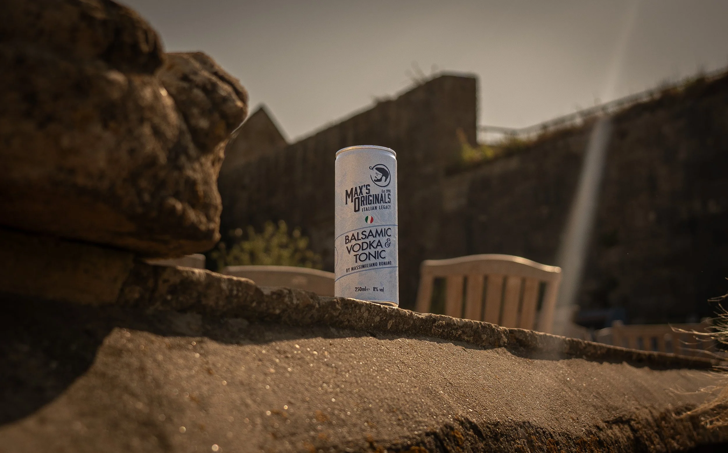 A can of Max's Originals Balsamic Vodka & Tonic positioned on a ledge outdoors in front of a stone wall, with chairs and a wooden fence in the background, under sunset lighting.