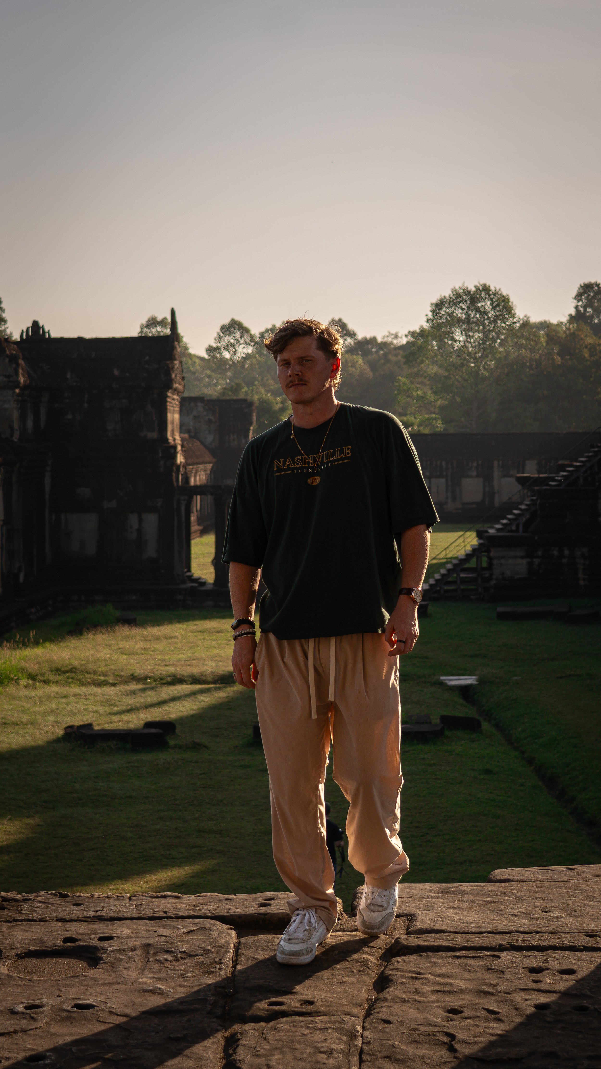 A young man walking on a stone path with ancient ruins and trees in the background during sunset.
