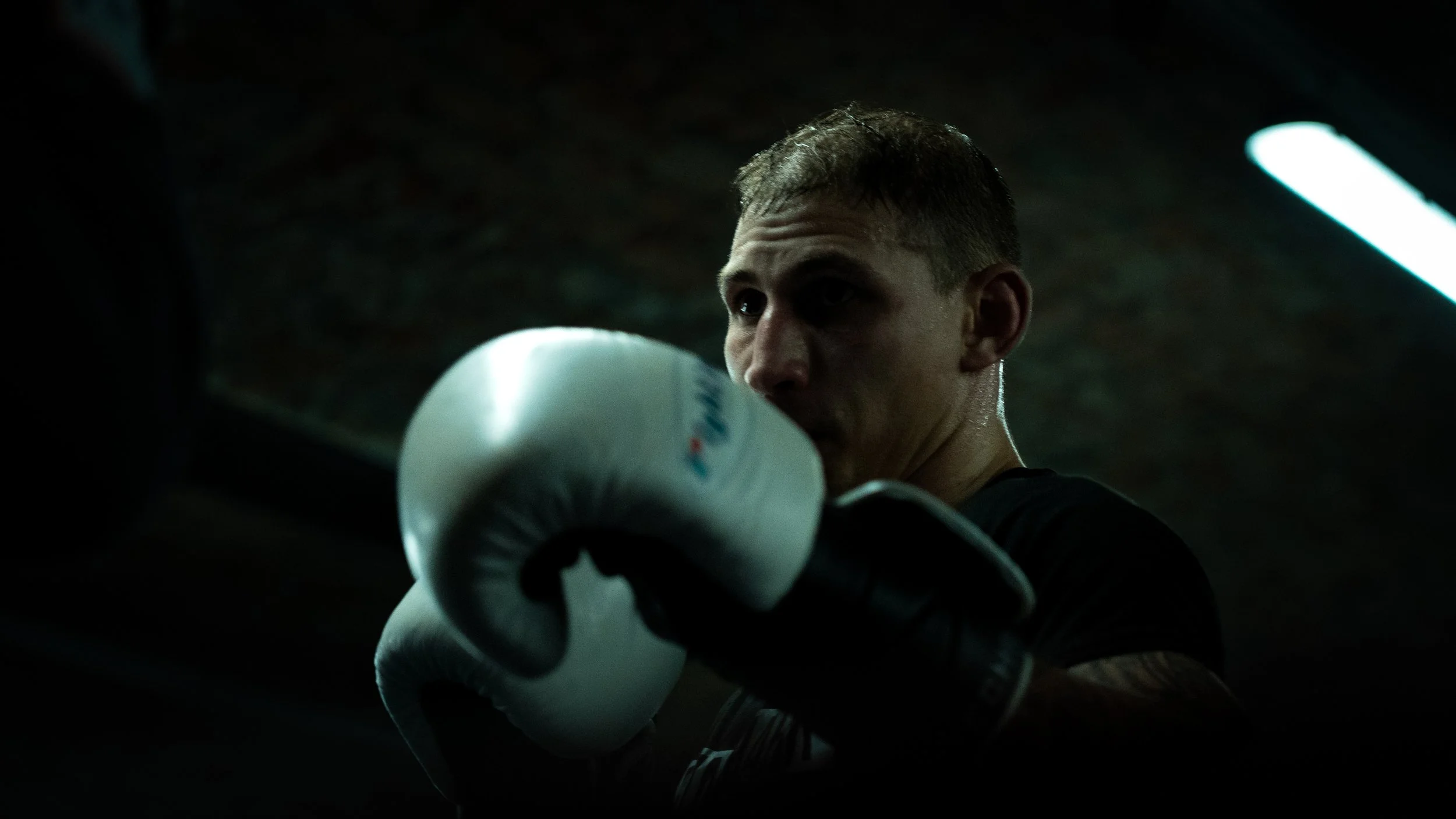 A boxer wearing black gloves is throwing a punch in a dimly lit gym, with a focused expression on his face.