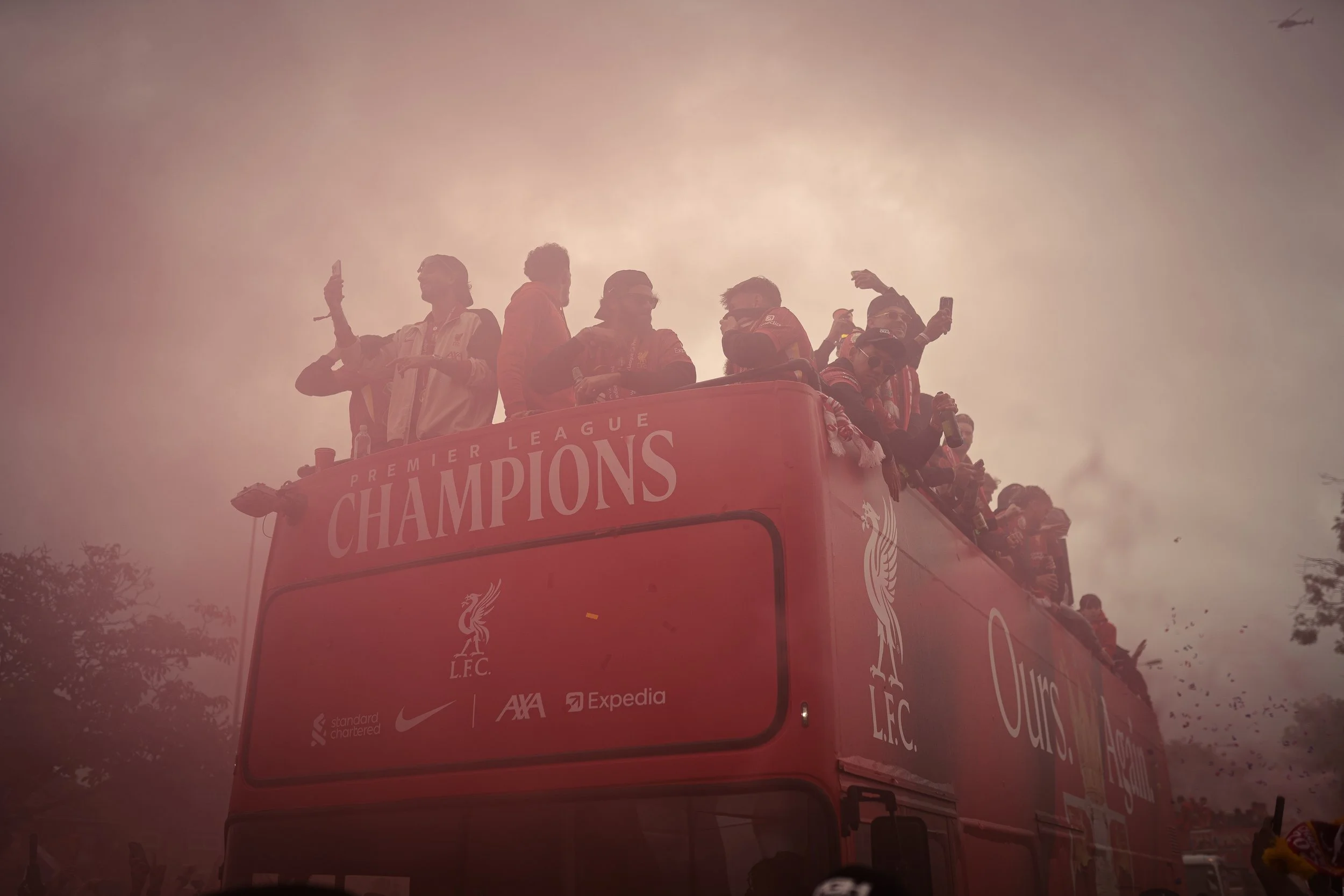 Liverpool FC players celebrating on a bus with a banner reading 'Premier League Champions' during the daytime, with a cloudy sky and some trees in the background.