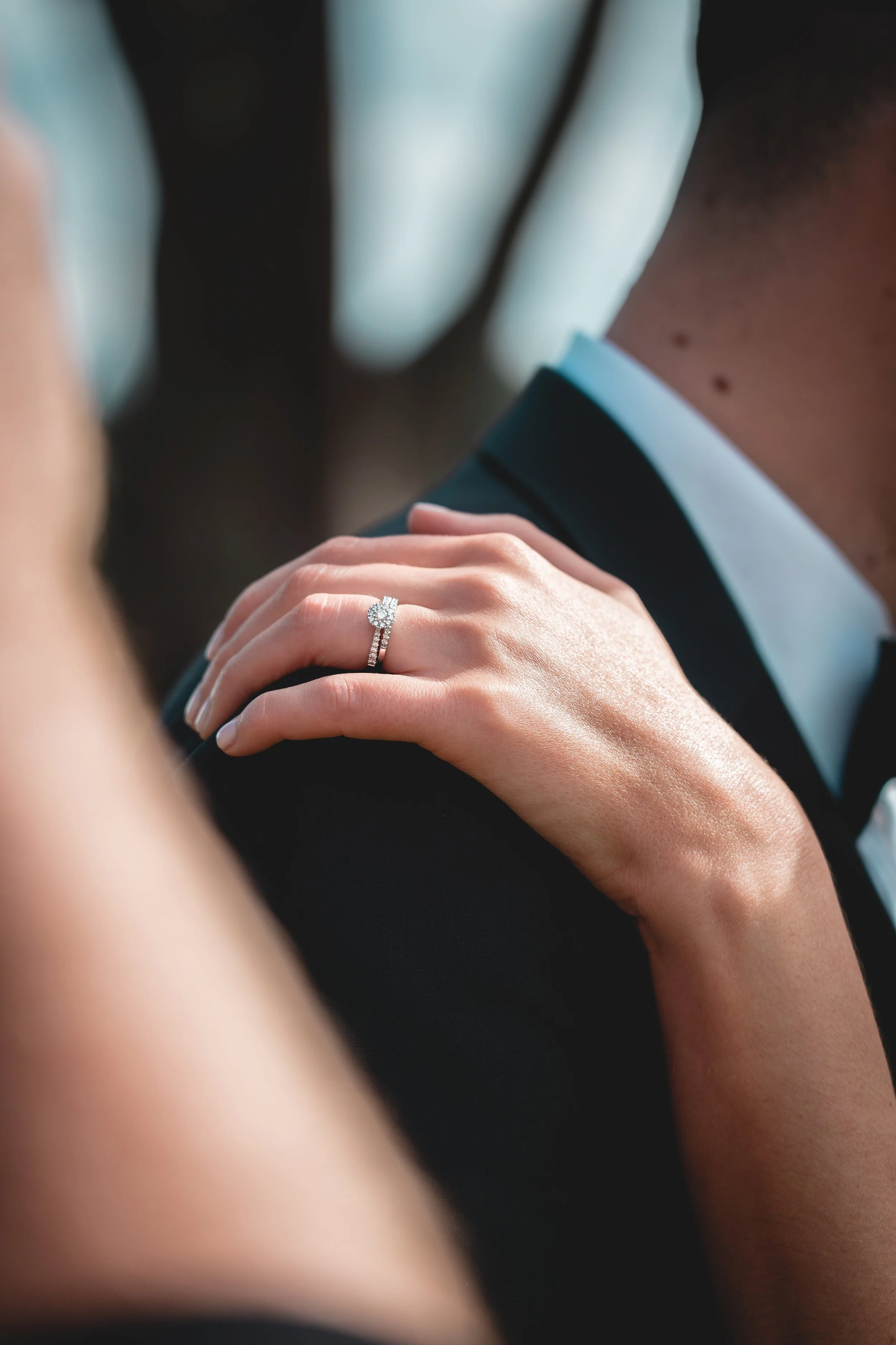 Close-up of a woman's hand wearing an engagement ring and wedding band, resting on a man's shoulder, with a blurred background.