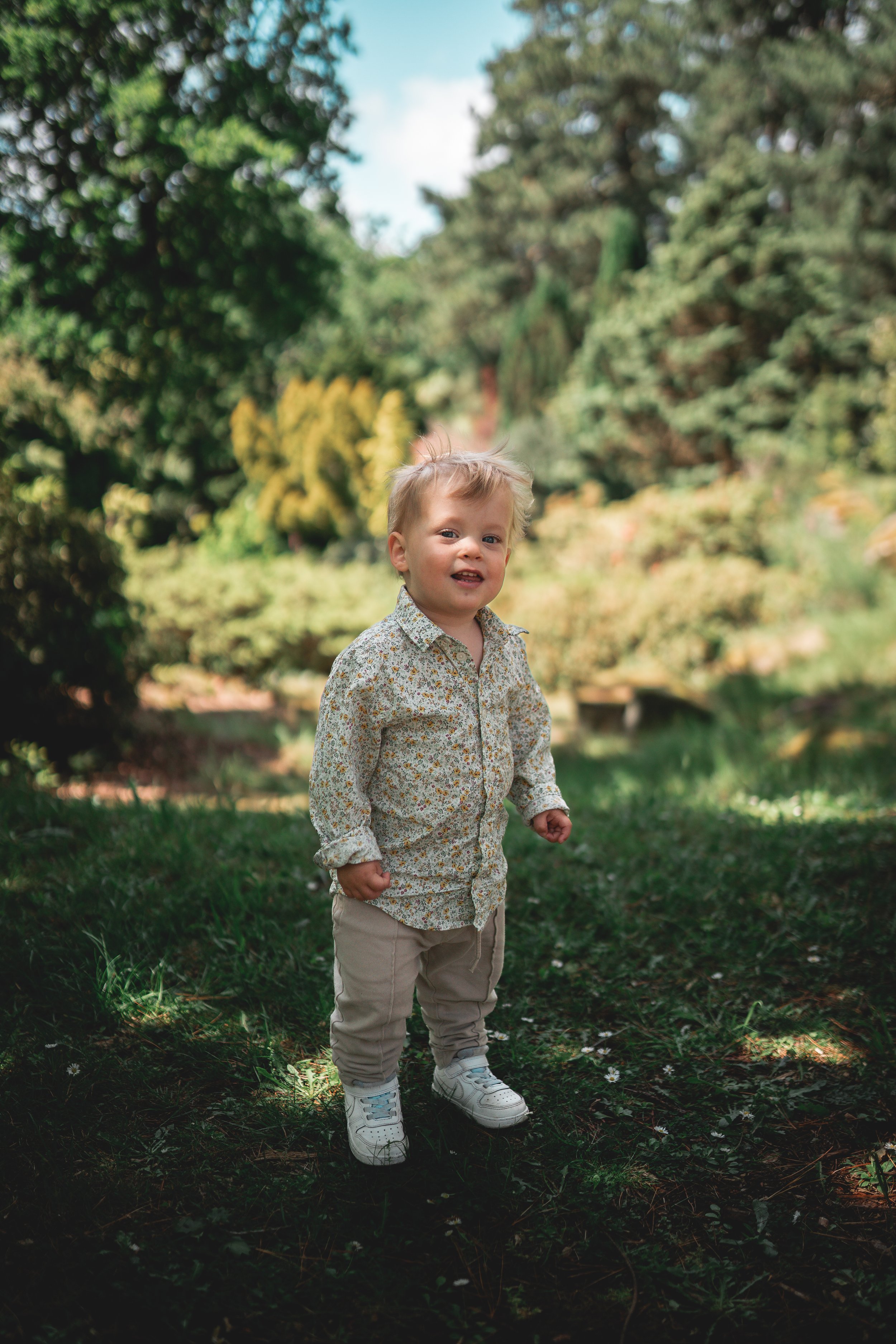 A young child standing outdoors in a lush green park or garden, smiling at the camera. The child is dressed in a long-sleeve, floral-patterned shirt, beige pants, and white sneakers. Tall trees and bushes are visible in the background under a blue sk