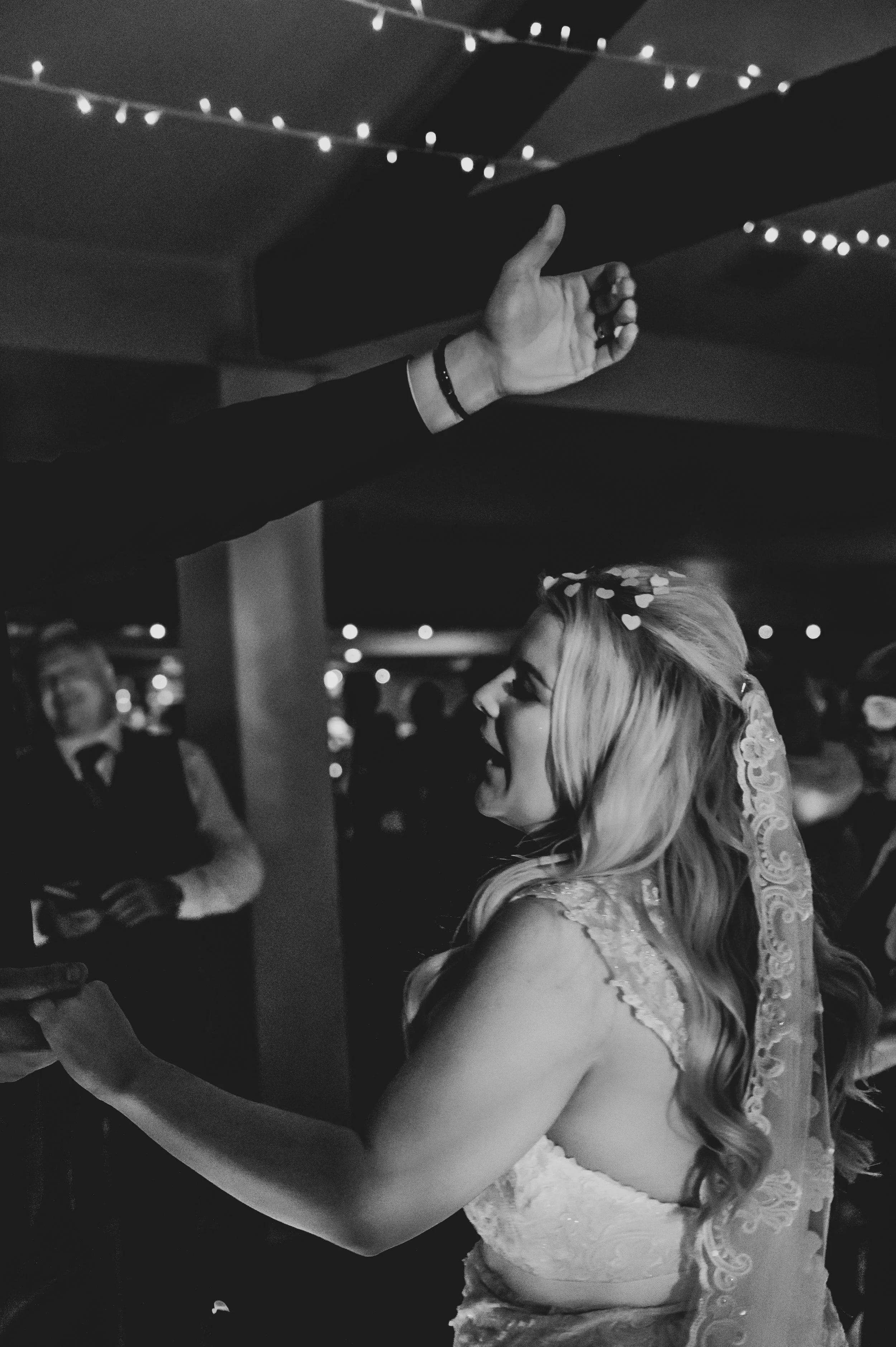 A bride with long blonde hair, wearing a lace wedding dress and veil, smiles and reaches her hand toward a raised arm in an indoor wedding reception with string lights overhead.