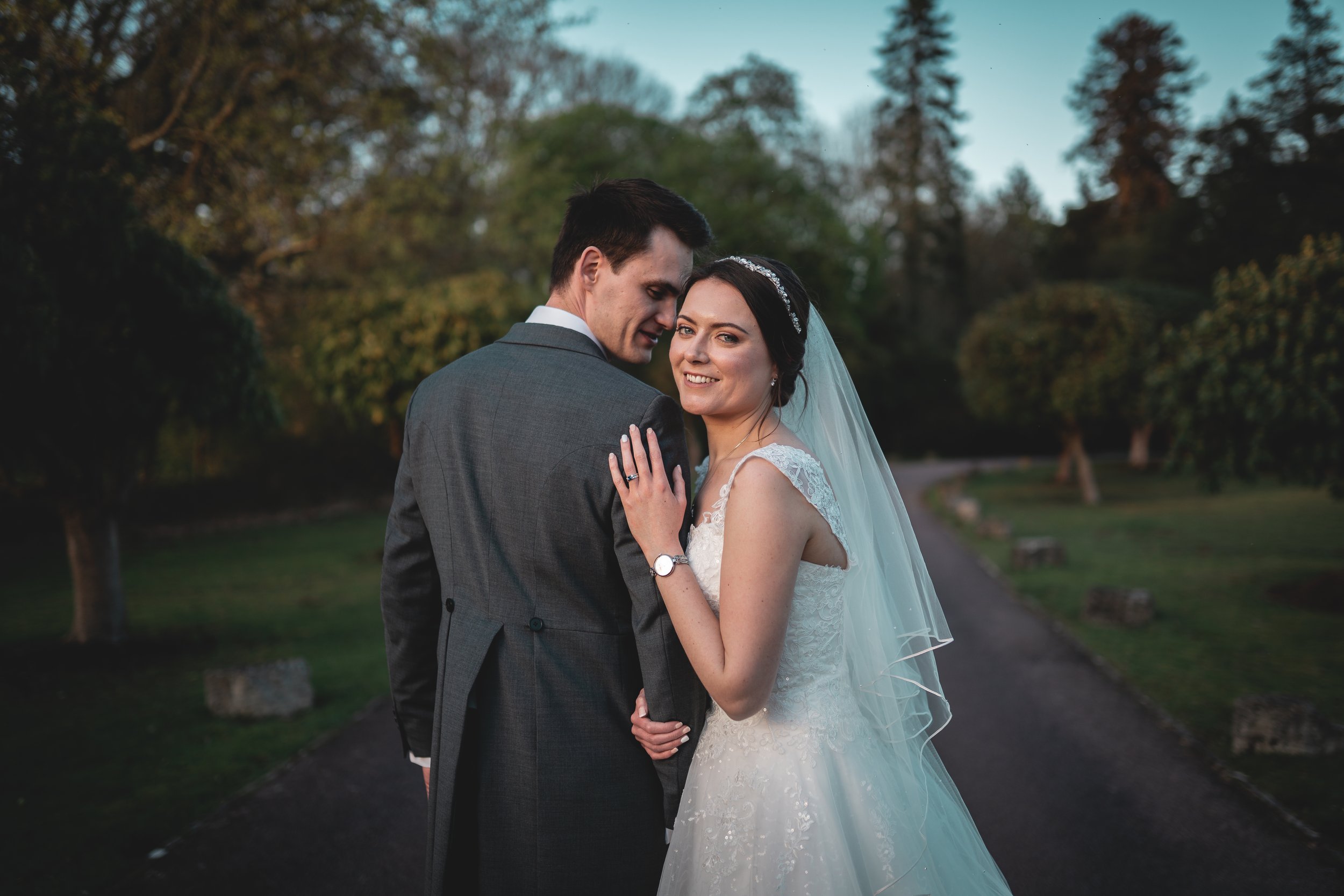 A bride and groom standing on a path amidst trees outdoors during sunset, smiling at the camera. The bride wears a white wedding dress with lace details and a veil, and the groom wears a dark gray suit.