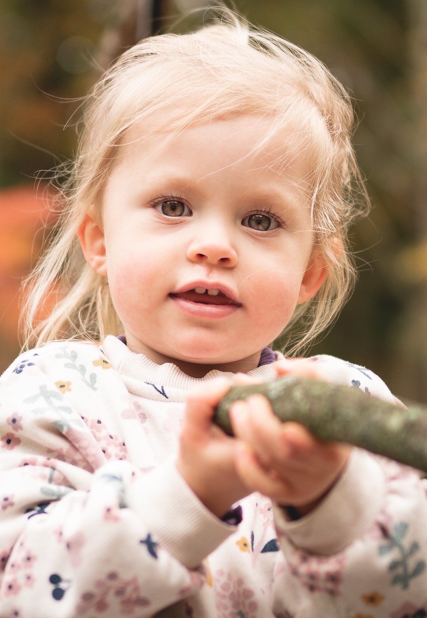 A young girl holding a stick outdoors with fall foliage in the background.