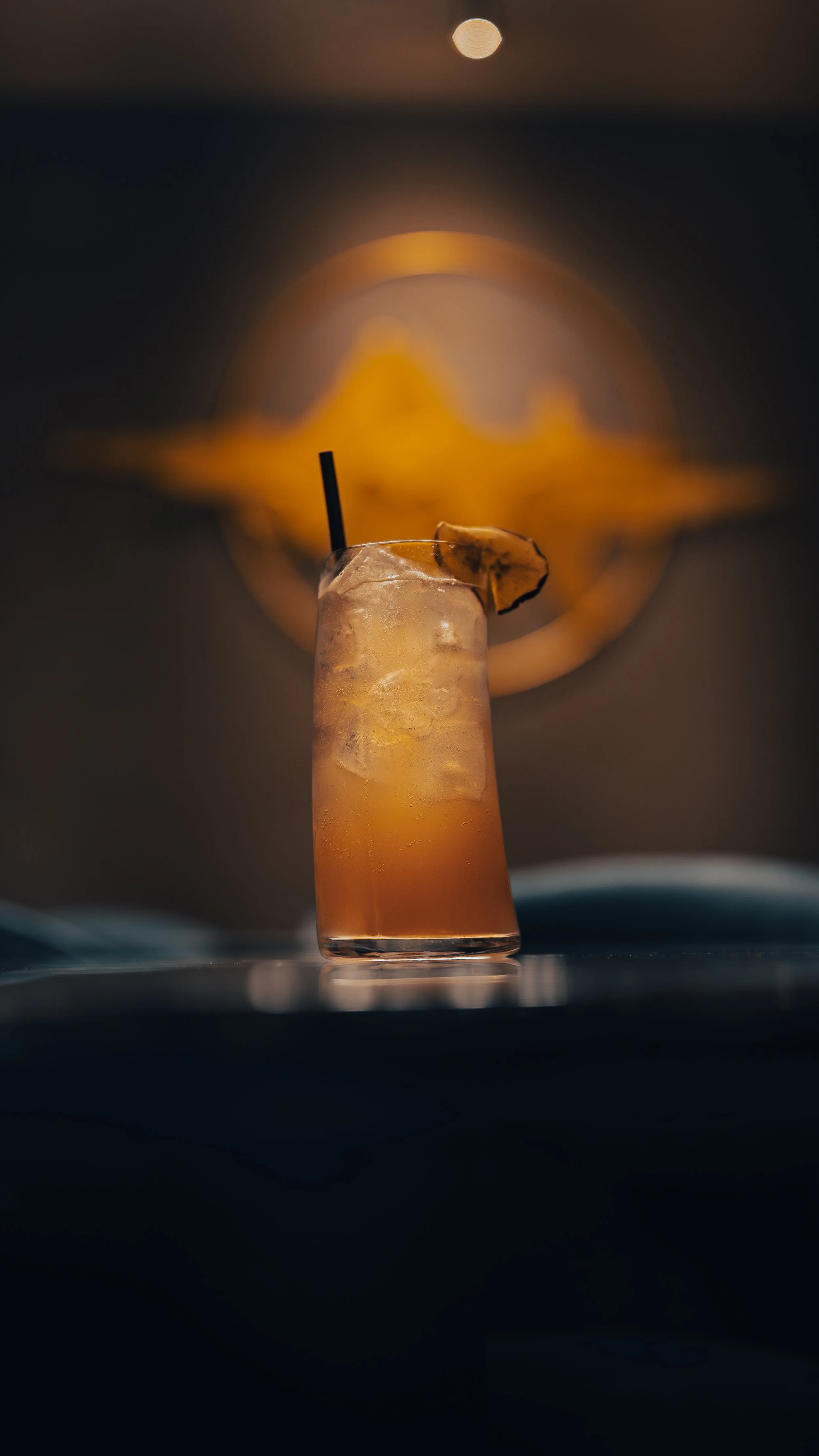 A tall glass of iced cocktail with ice cubes and a lemon slice garnish, set against a dark background with a blurred logo