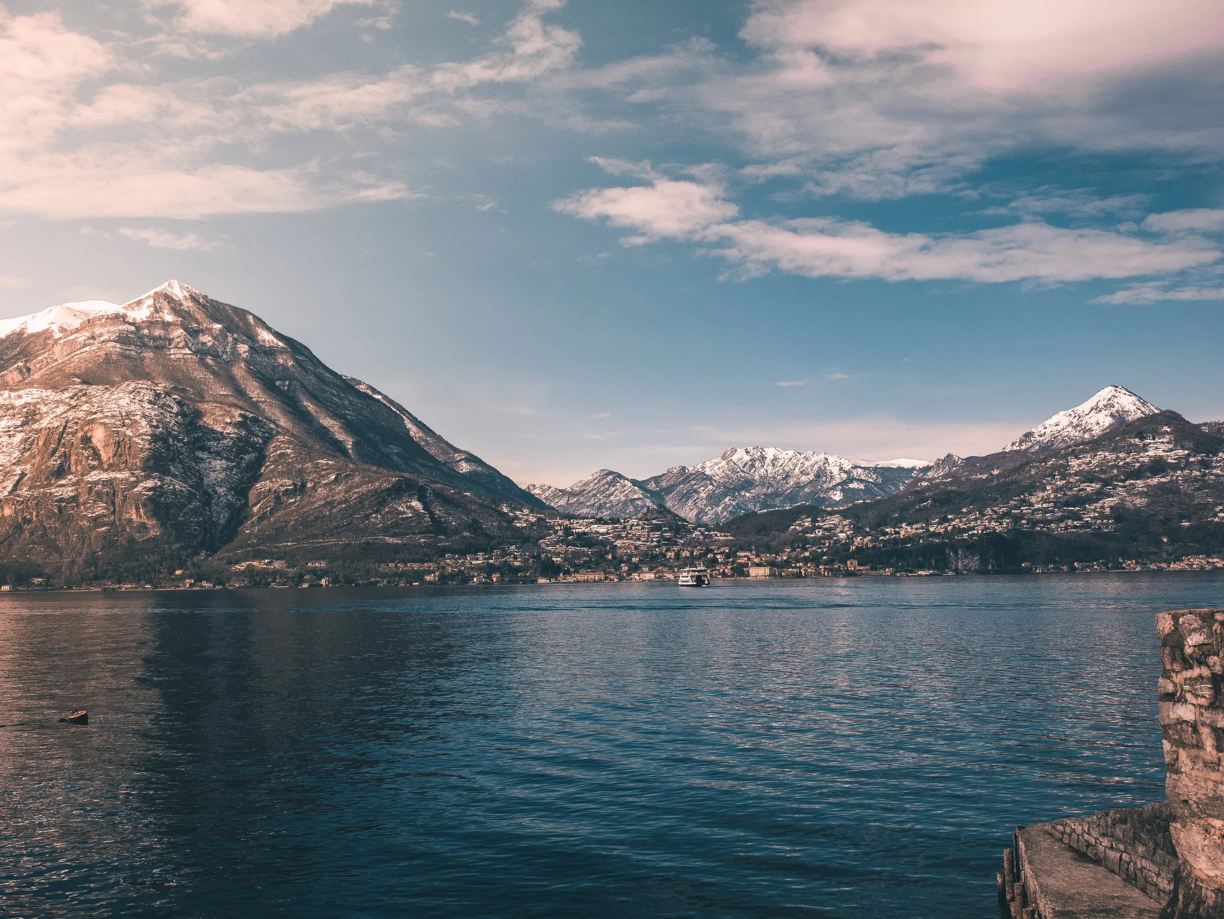Snow-capped mountains near a large body of water with a small boat, under a partly cloudy sky.