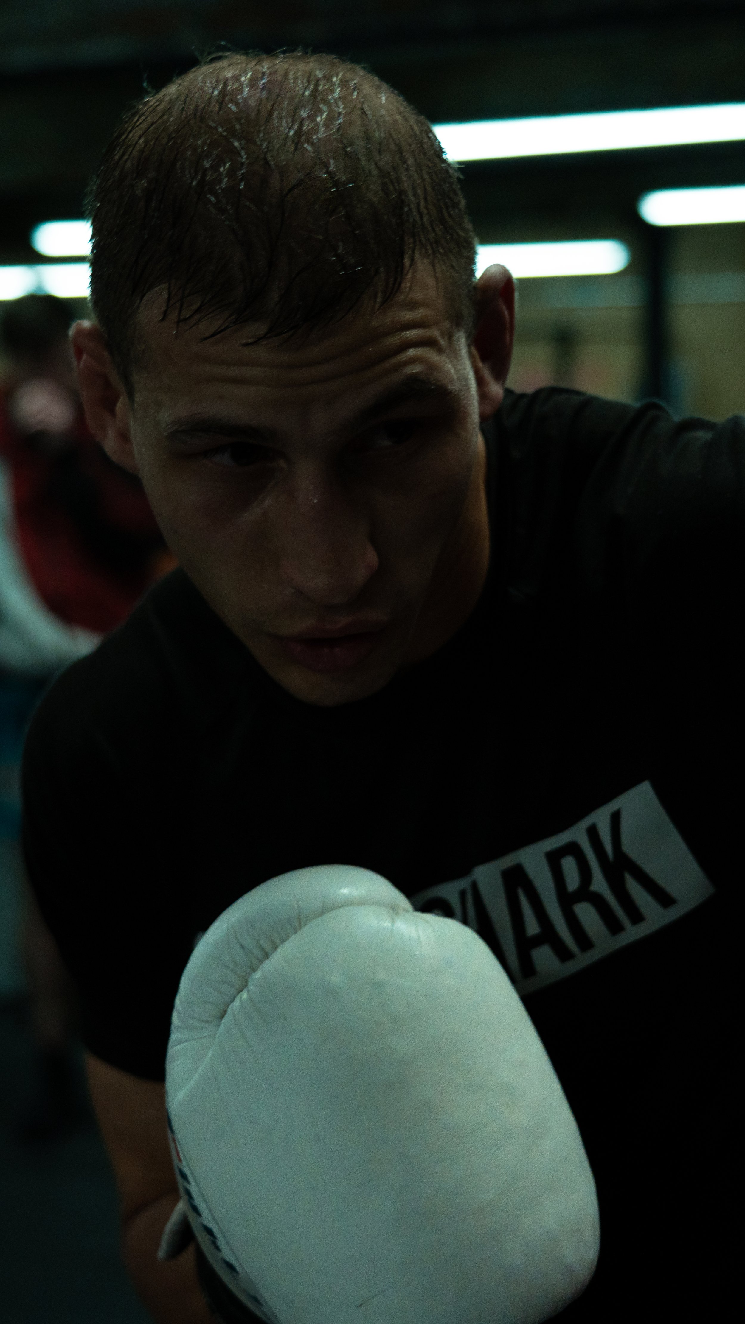 A male boxer with wet hair wearing a black shirt and white boxing gloves, in a boxing gym, with another person blurred in the background.
