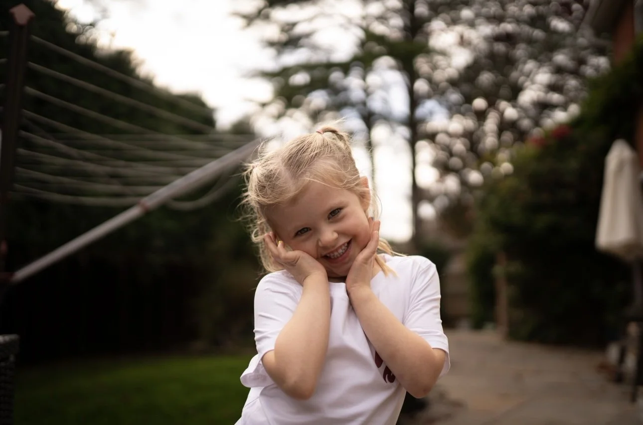 A young girl with blonde hair tied in a ponytail, smiling and posing with her hands on her cheeks outdoors in a garden or backyard.