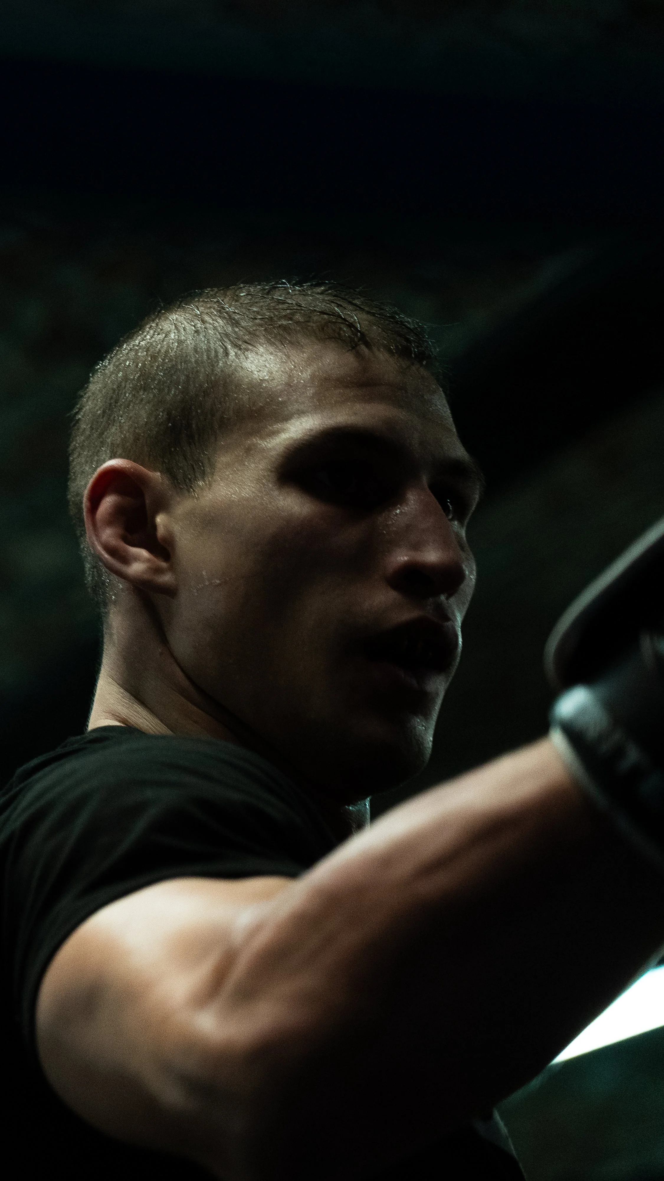 A young man with short hair and sweat on his face, gripping a camera or equipment, in a dimly lit environment.