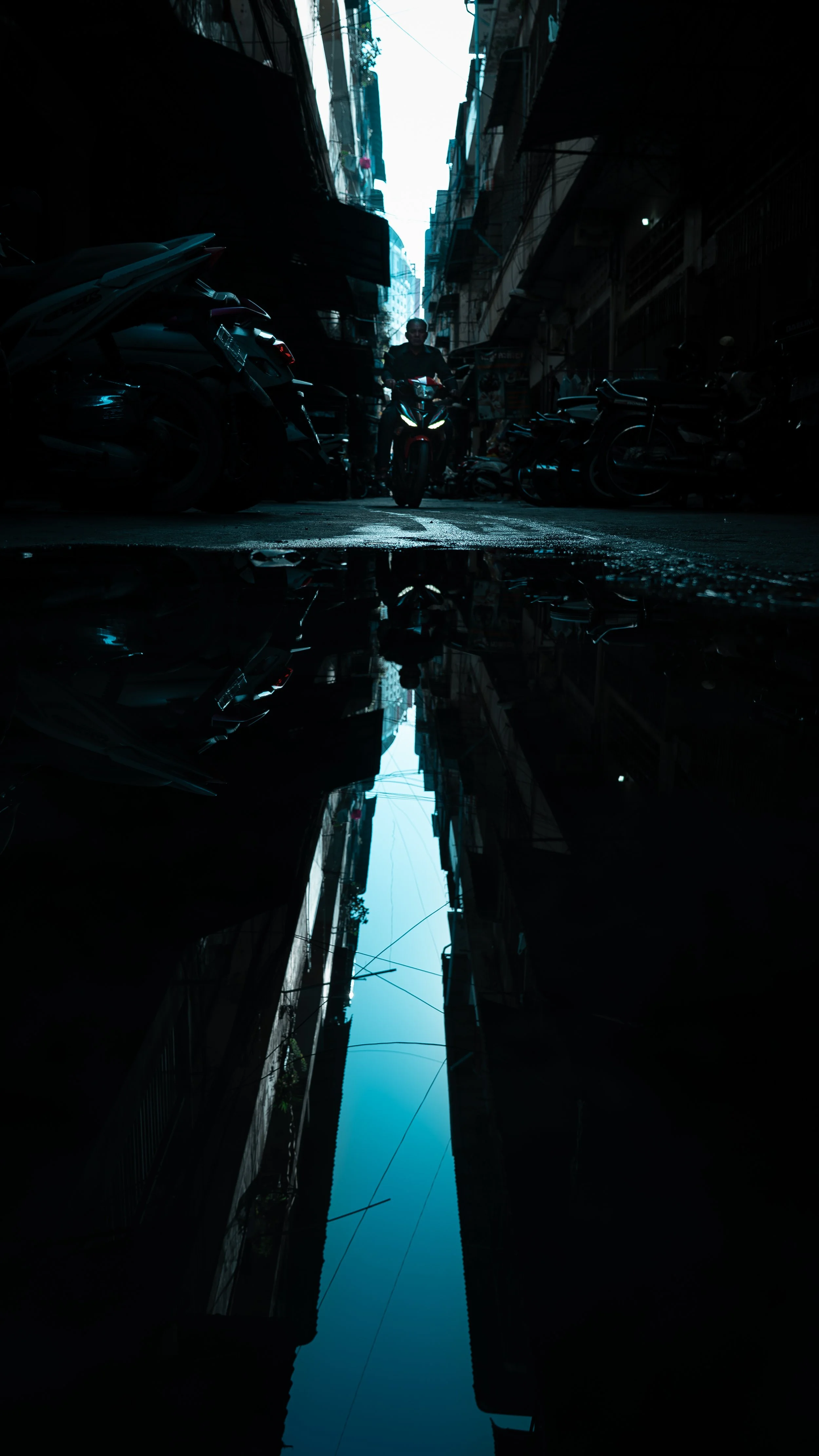 A dark alleyway with parked motorcycles and bicycles, a person riding a scooter, and a puddle reflecting the sky and buildings above.