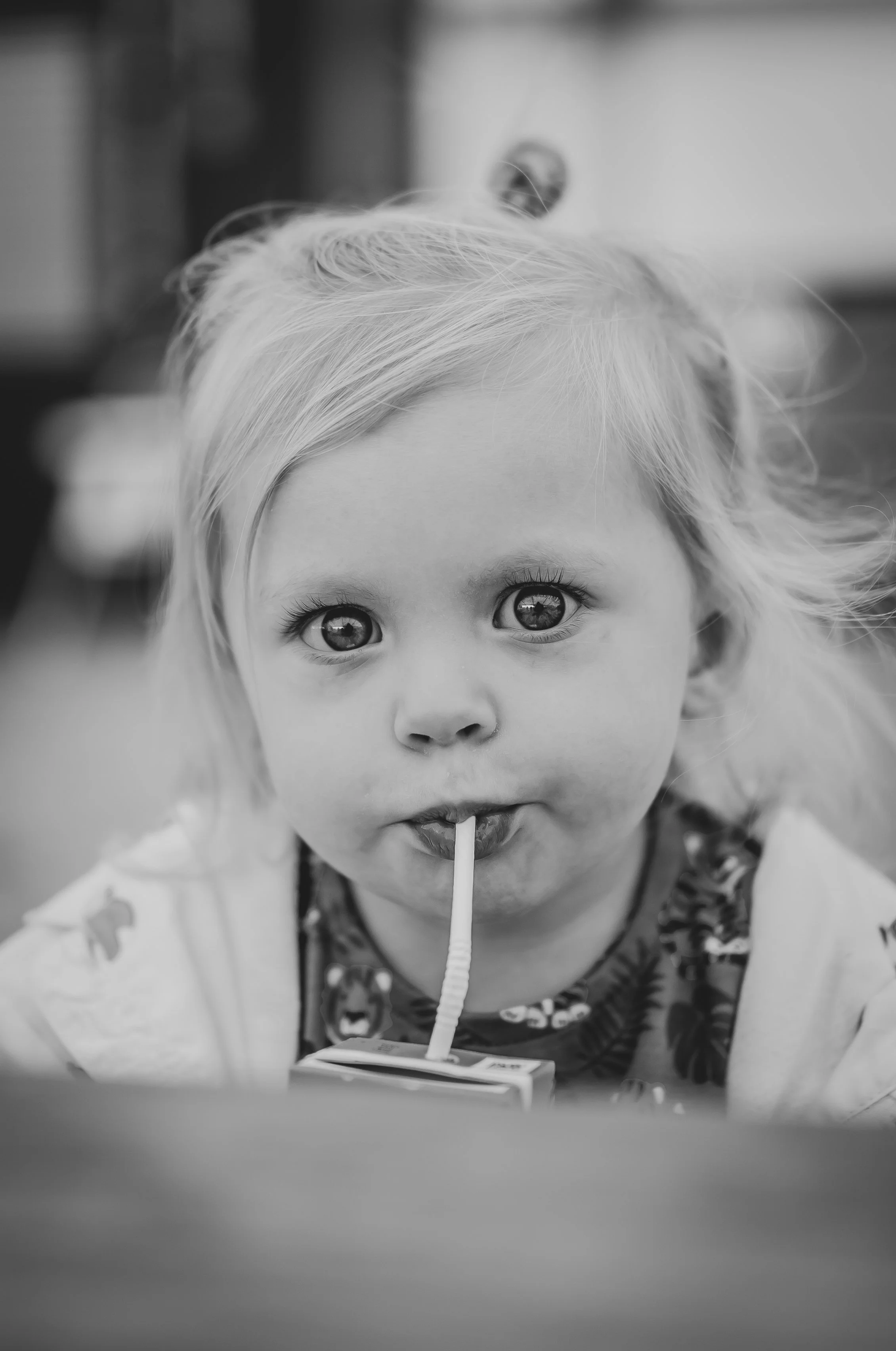 A young girl with light hair drinking from a juice box with a straw, looking directly at the camera. Black and white photograph.