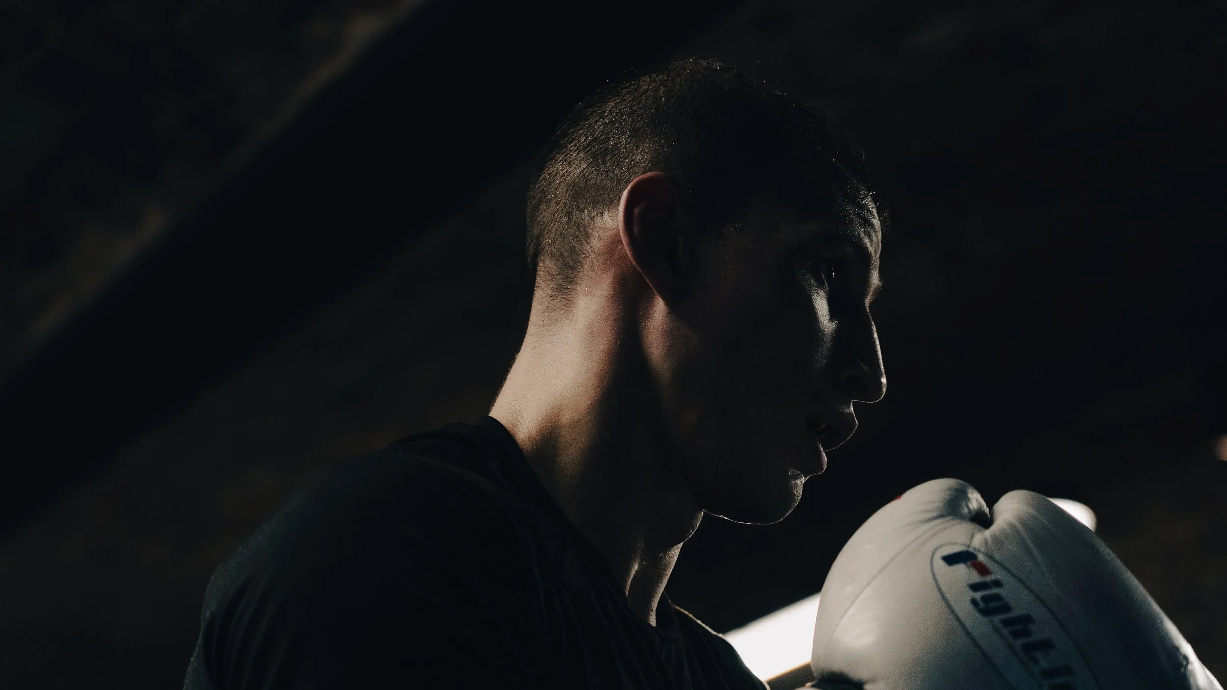A male boxer in a dark environment, wearing boxing gloves, with a serious expression.