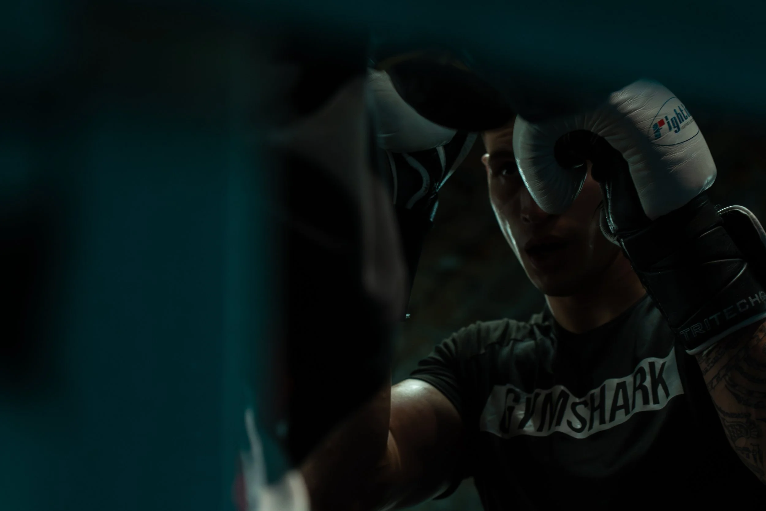 A woman in boxing gear, wearing gloves and a GUNSHARK T-shirt, in a gym or boxing ring, focusing during training.