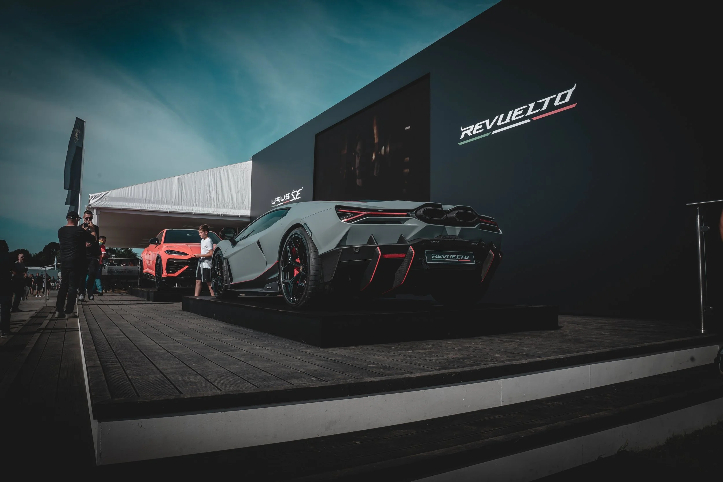 A display of two high-performance sports cars on a raised platform at an automotive event, with a large black backdrop featuring the word REVUELTO. The cars include a matte gray Lamborghini with red accents and a red car behind it. Several people are