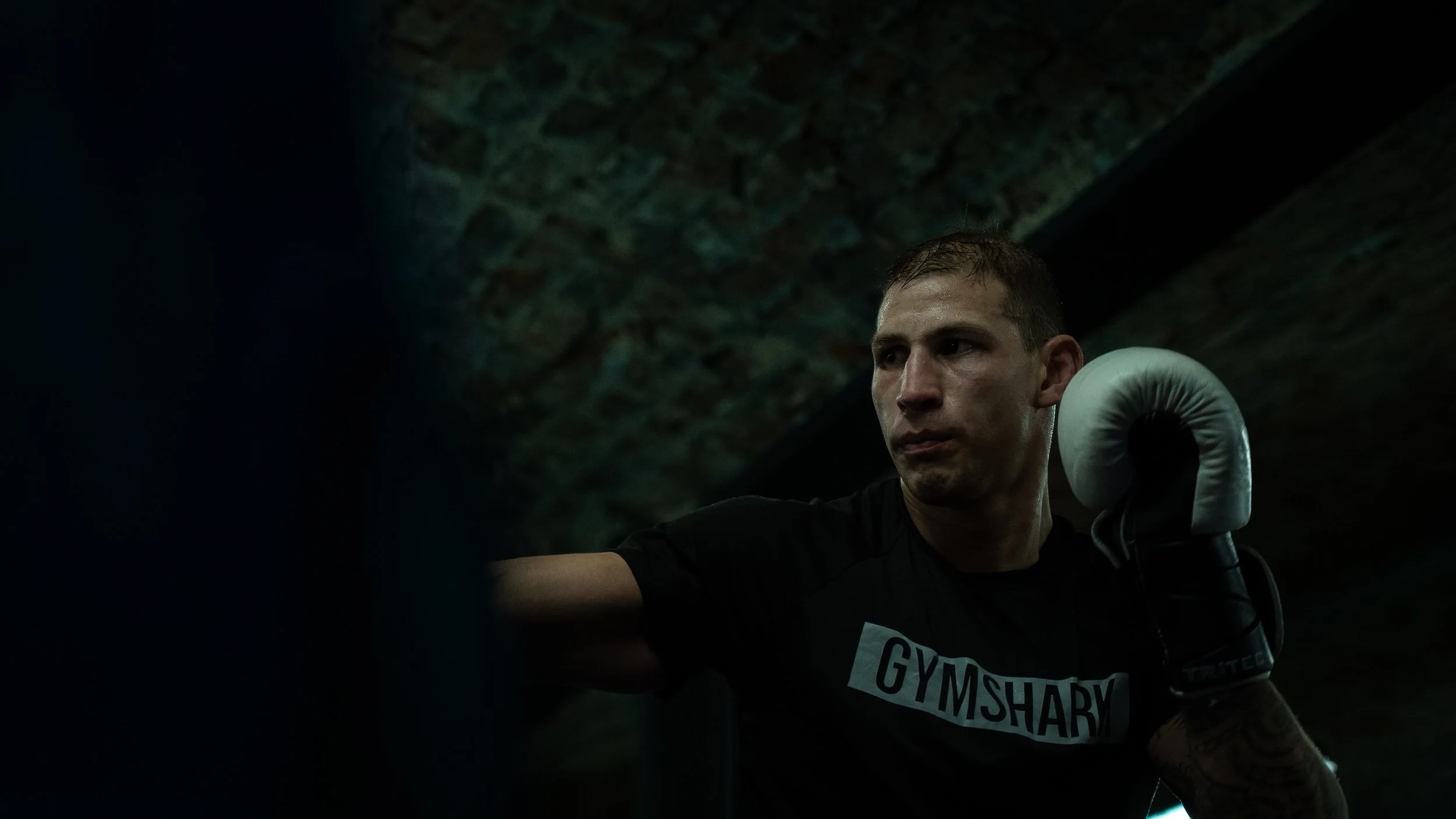 A man in a black t-shirt with 'GYMSHARK' written on it, wearing boxing gloves, looks focused during a workout in a dark, industrial-style gym with exposed brick walls.