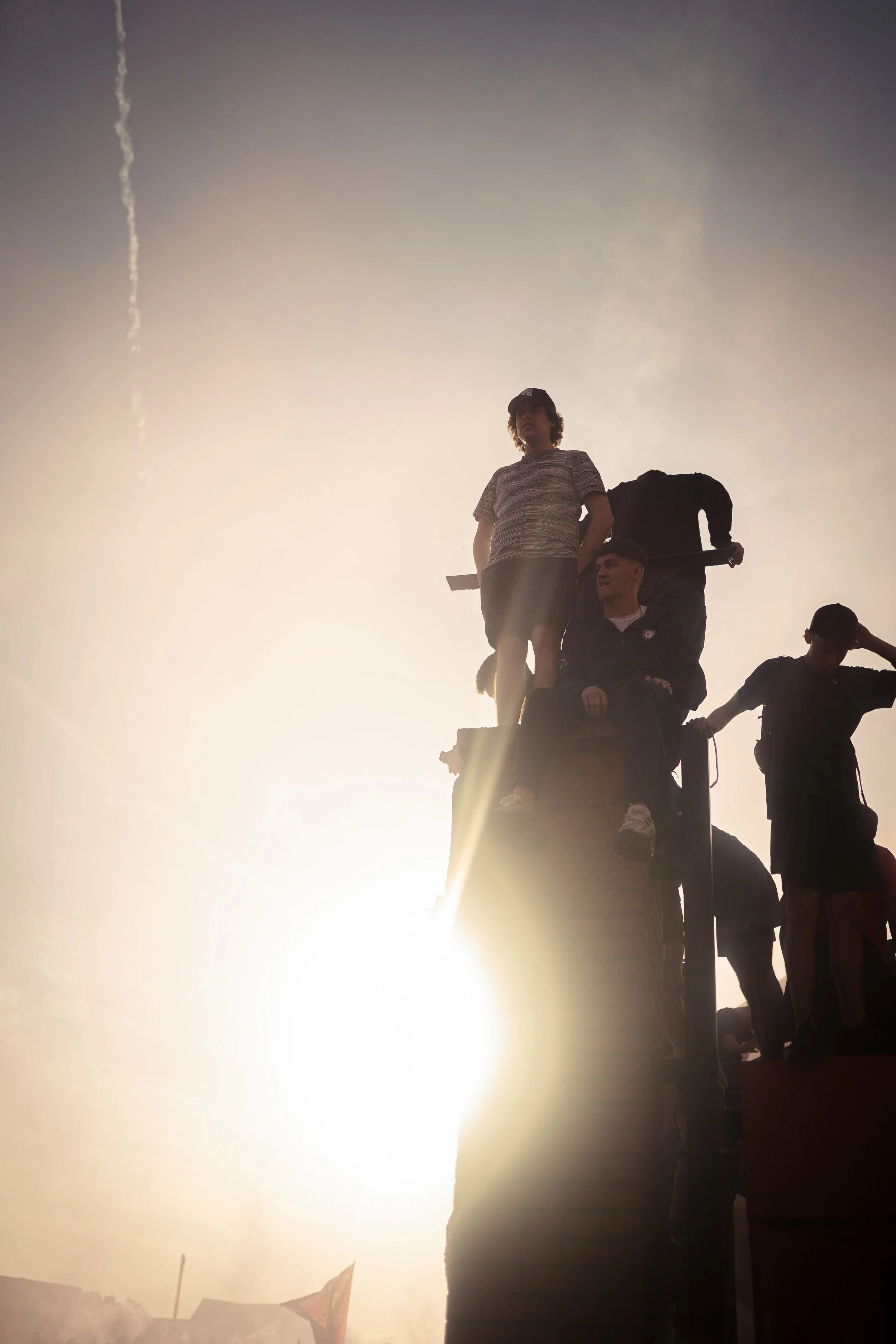 People sitting on a structure during a sunset with the sun behind them, creating a silhouette effect.