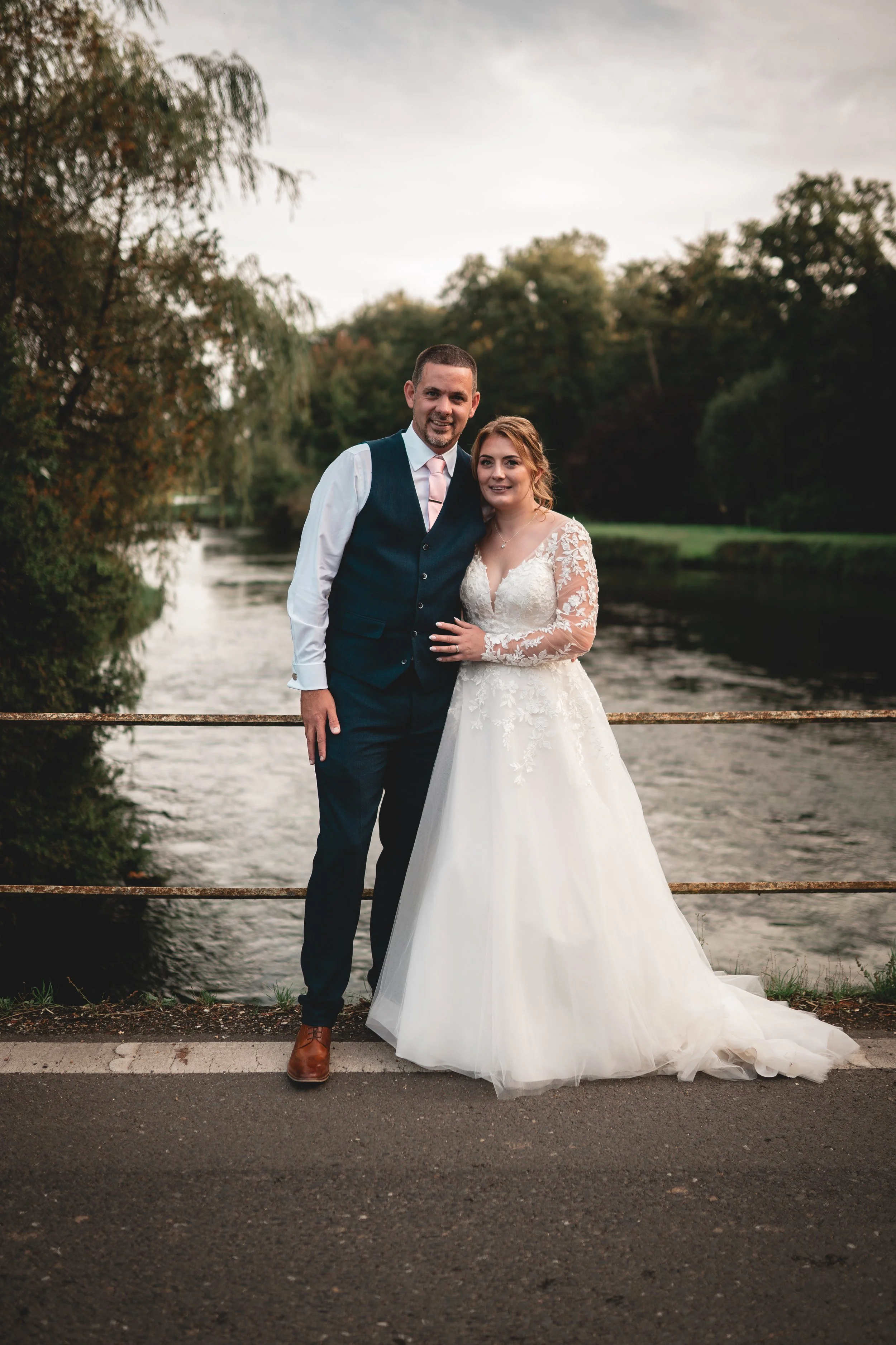 A bride and groom standing together on a bridge by a river, with trees and cloudy sky in the background, celebrating their wedding day.