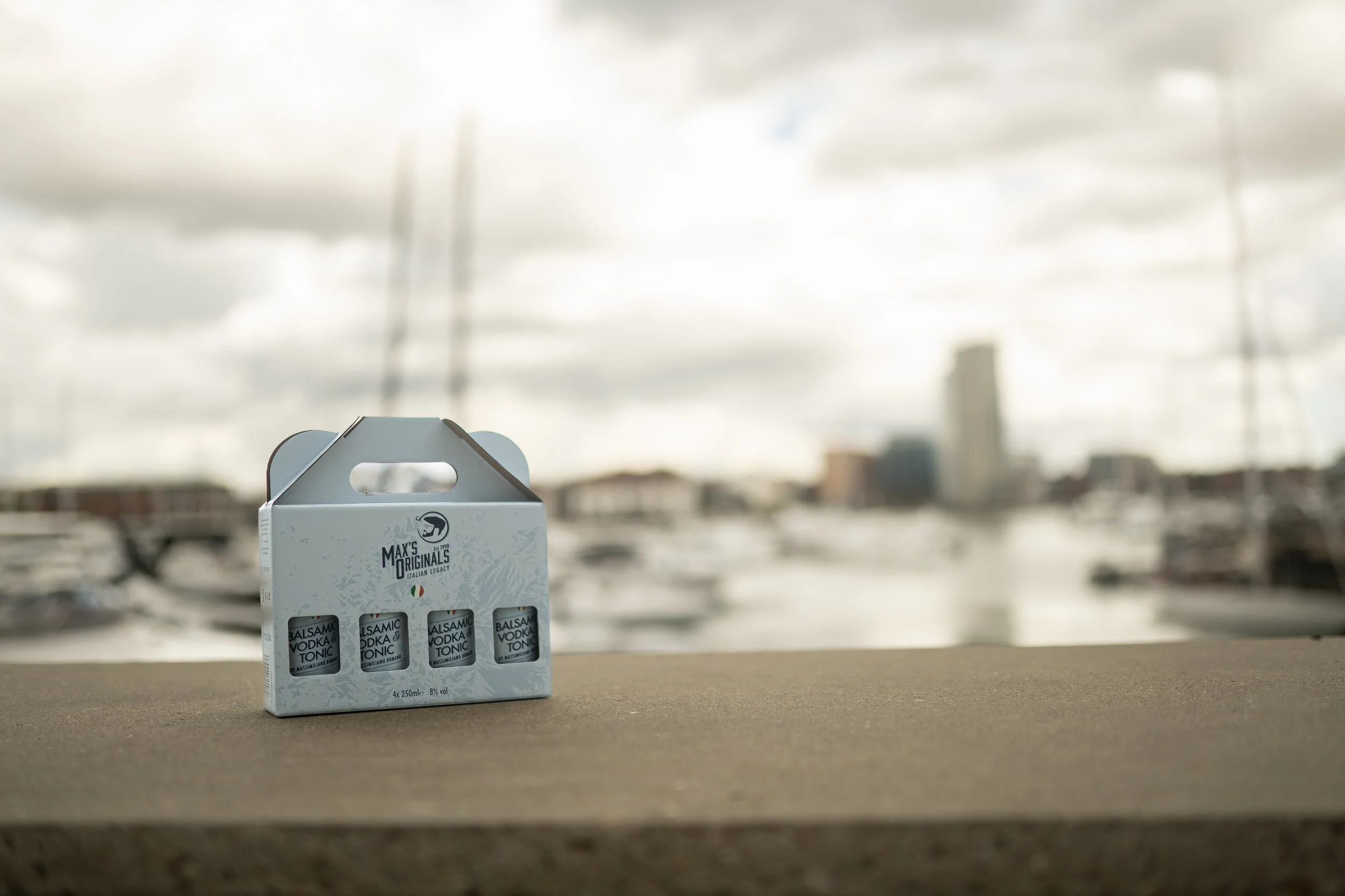 A small white gift box with four small bottles labeled "Balsamic Vodka Tonic" sits on a ledge near a marina, with boats and a city skyline in the blurry background.