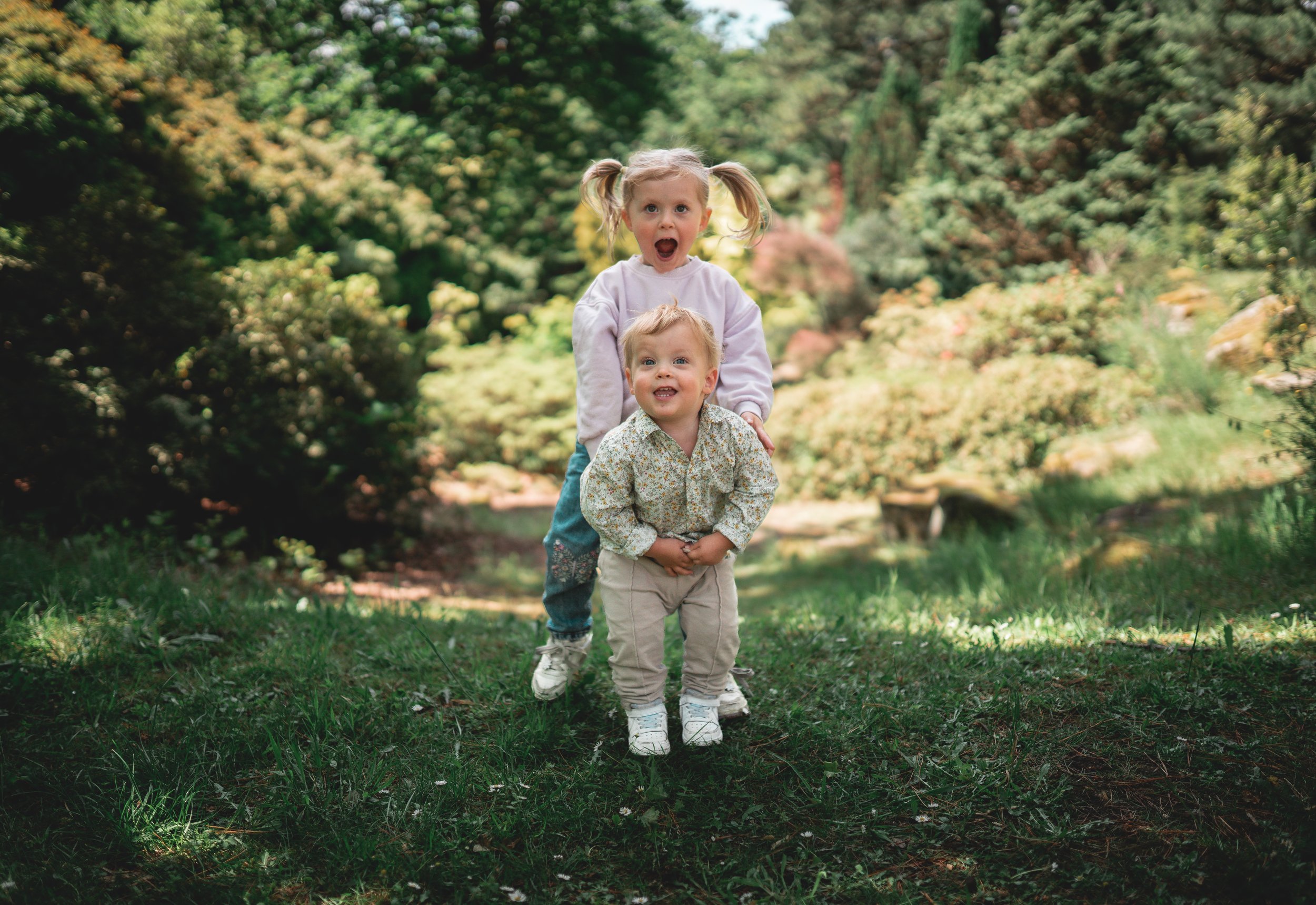 Two young children, a girl and a boy, playing outdoors in a lush, green garden or park with trees and bushes in the background. The girl is standing behind the boy, both smiling and looking toward the camera.