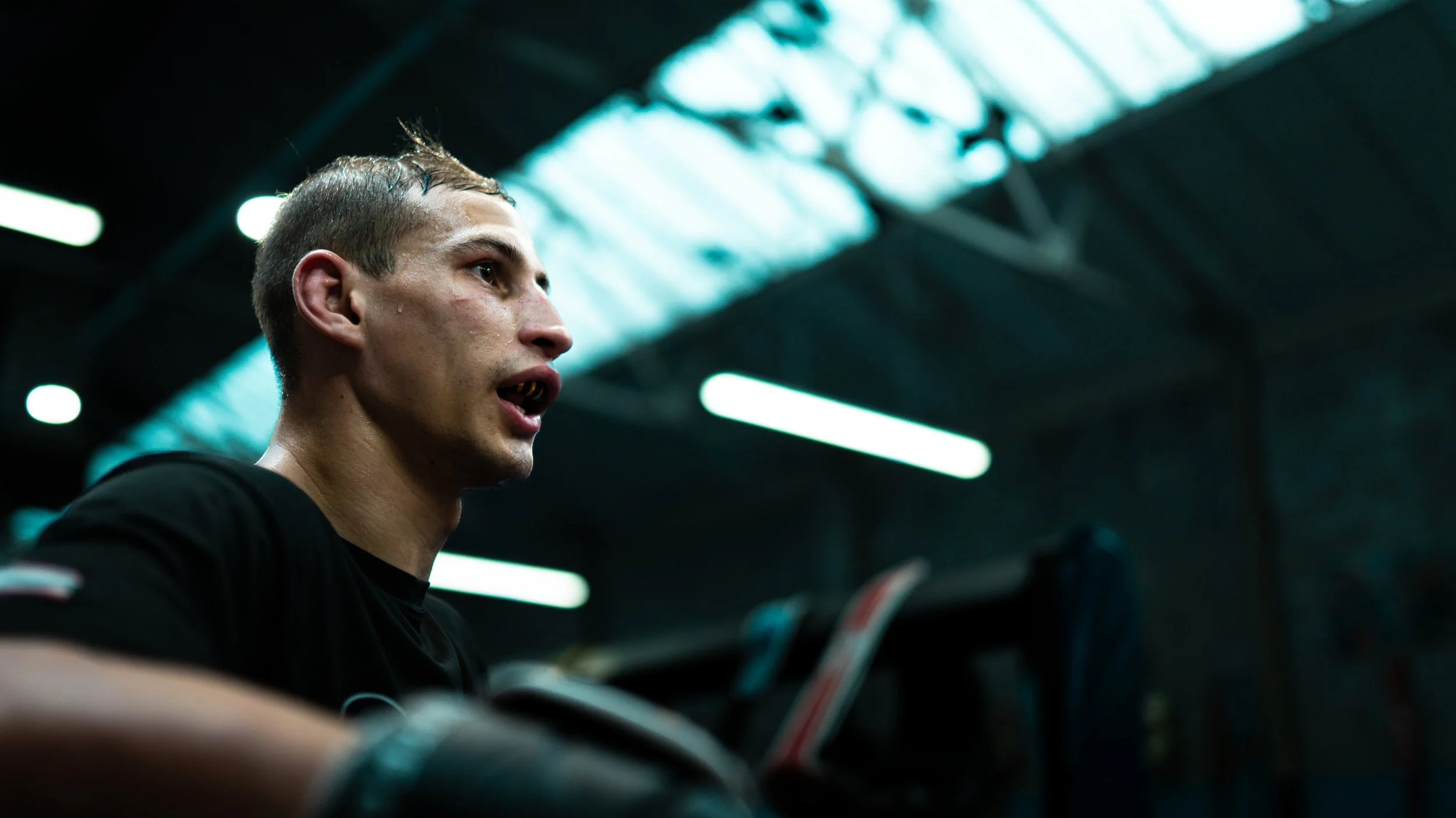A young man with sweaty face in a gym, lifting weights, with bright overhead lighting