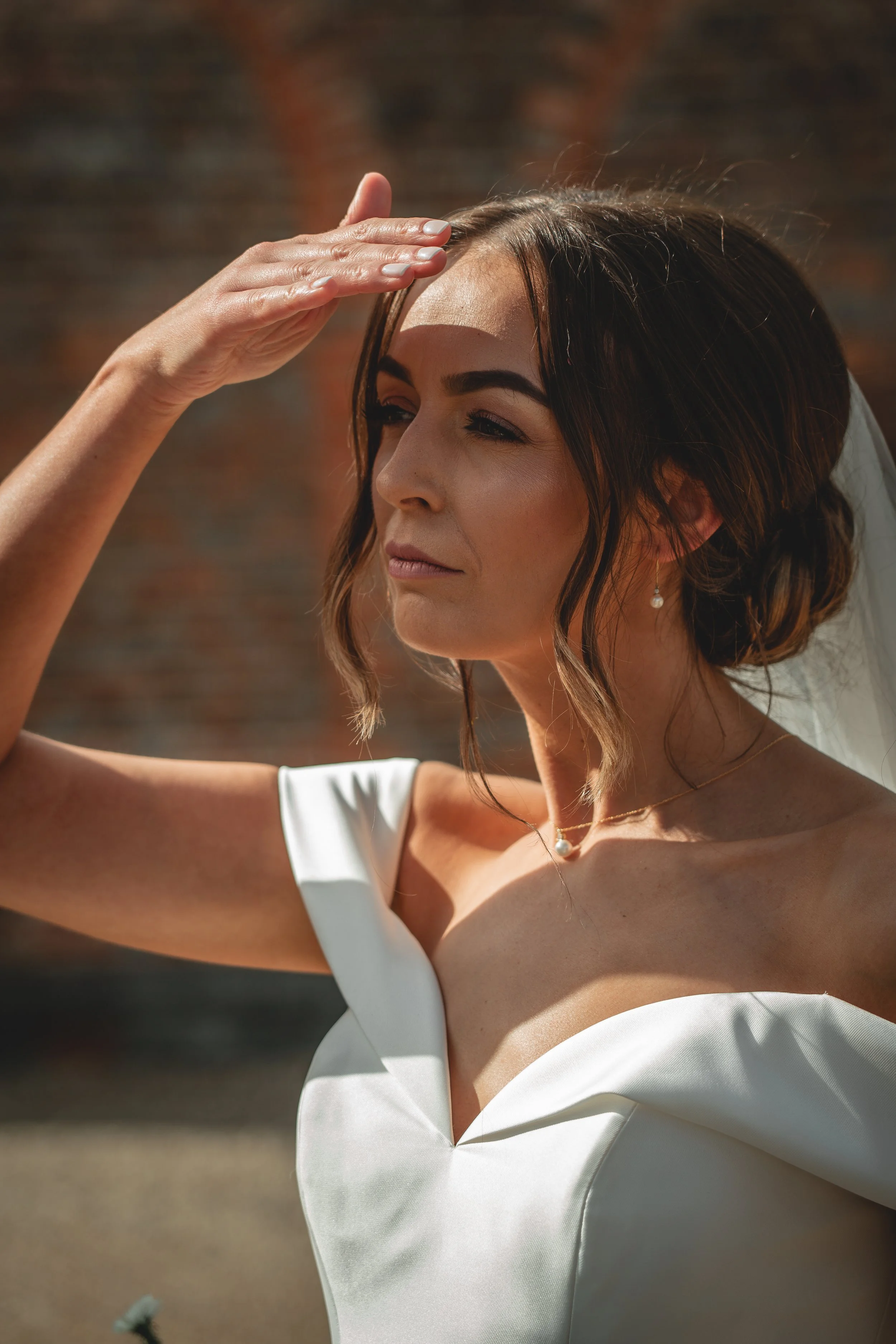 A woman in a wedding dress is shielding her eyes from the sunlight with her hand, looking into the distance.