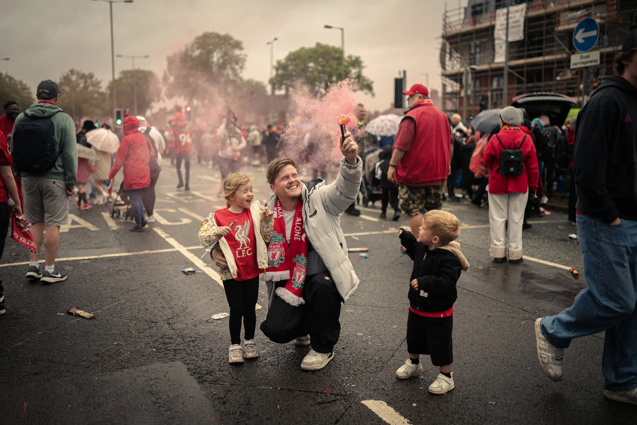 A lively street scene with a crowd celebrating, some wearing red and Liverpool FC scarves. In the foreground, a smiling man kneels with two children, holding a pink smoke flare, amid scattered debris and a smoky background.