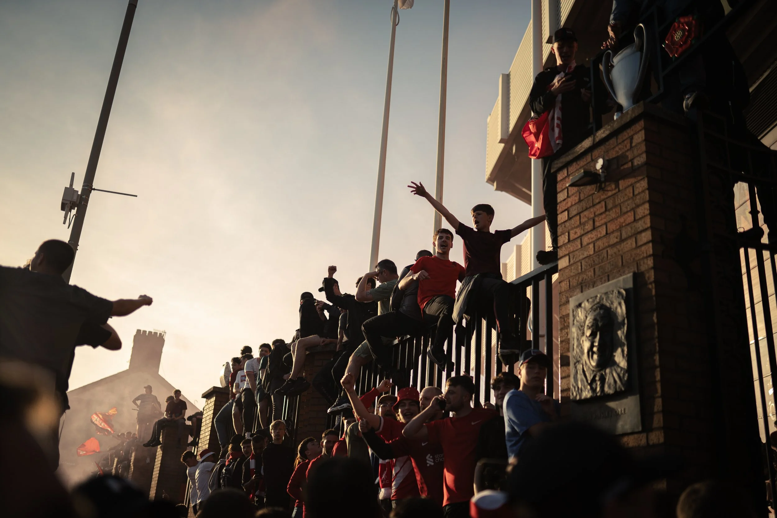 Group of young people gathered on a brick wall and railing, some sitting and some standing, celebrating and cheering during a sports event at sunset.