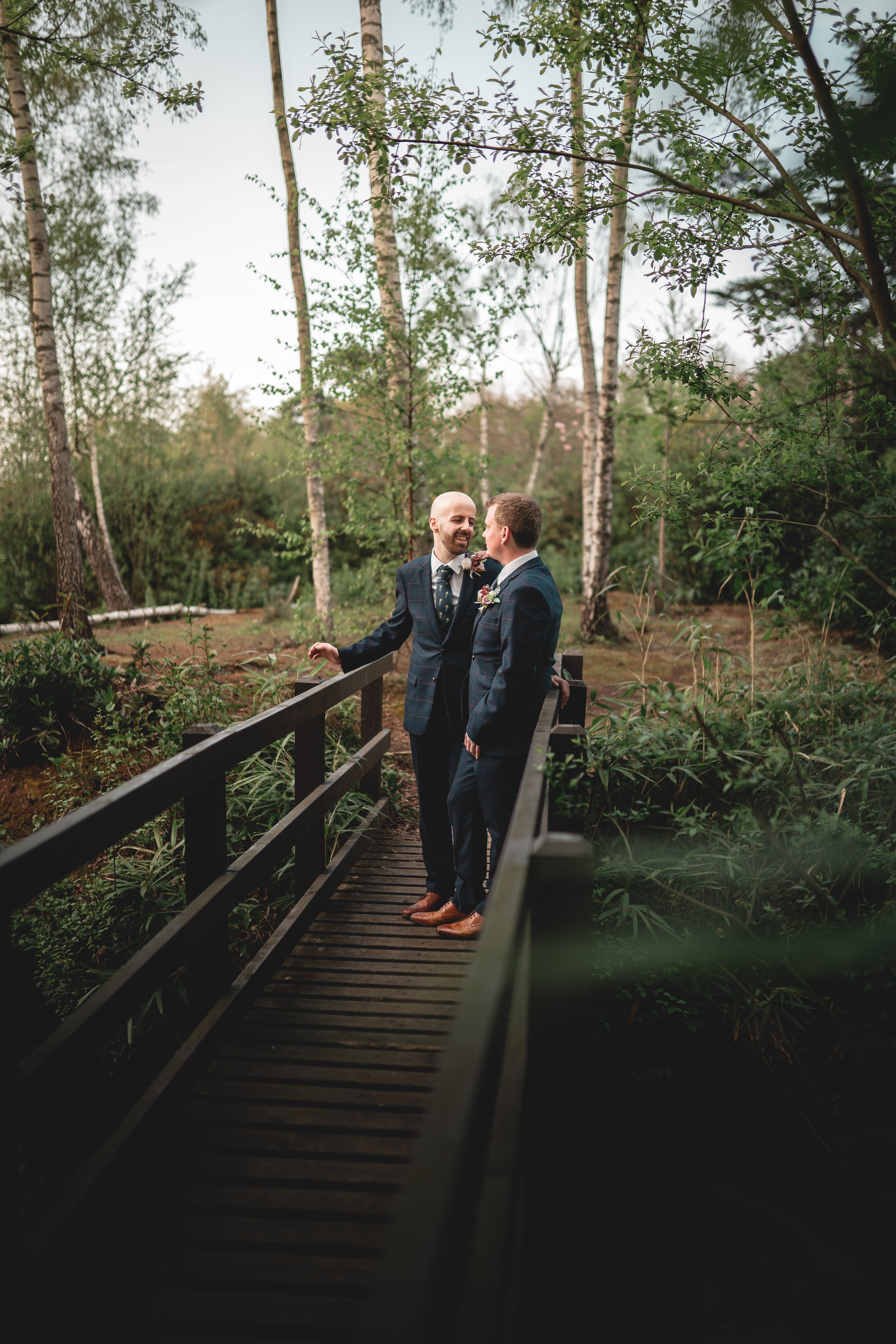 Two men dressed in suits standing on a small wooden bridge in a forest, smiling and looking at each other.