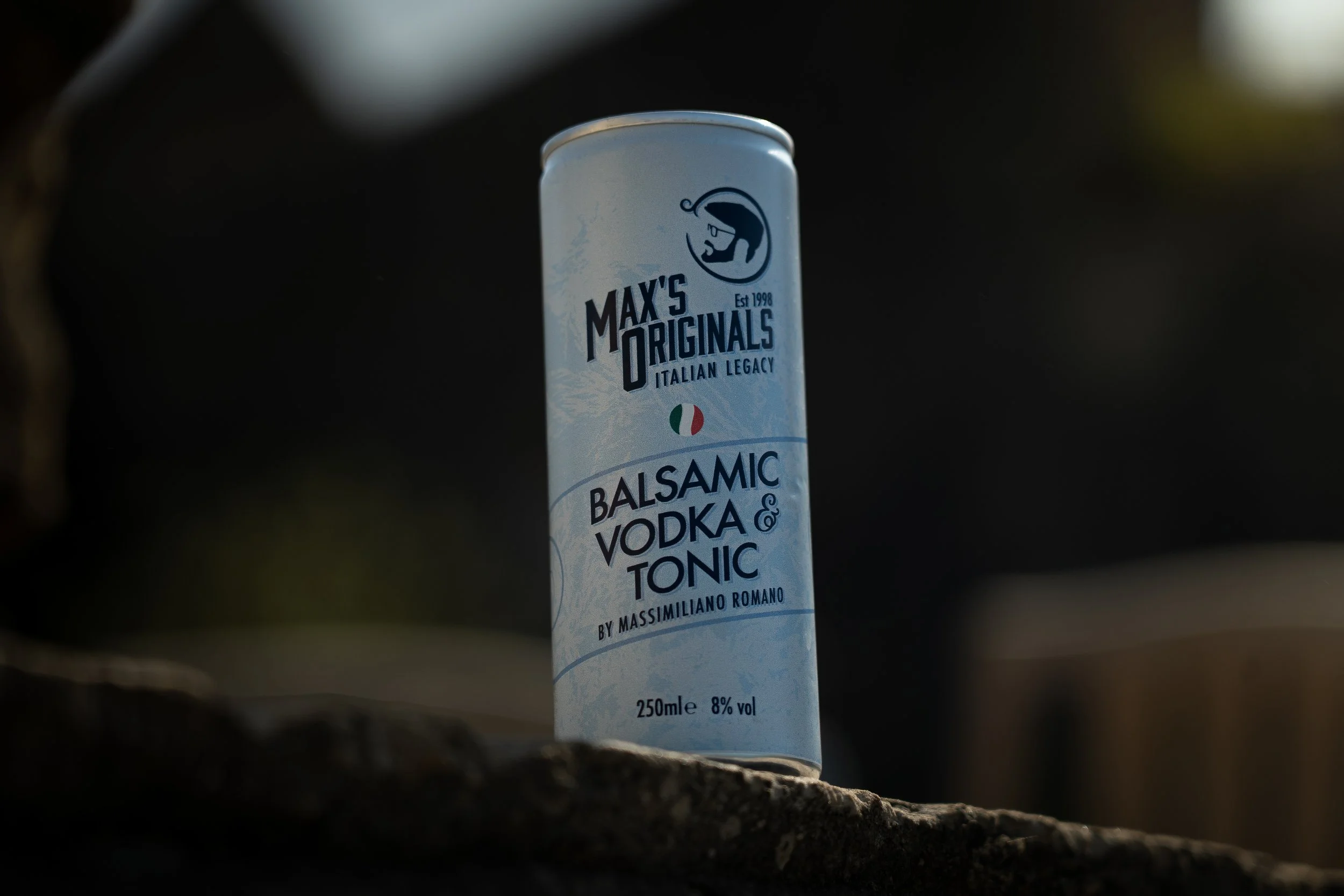 A tall, cylindrical can of Max's Originals Italian Balsamic Vodka & Tonic on a dark surface with a blurred background.