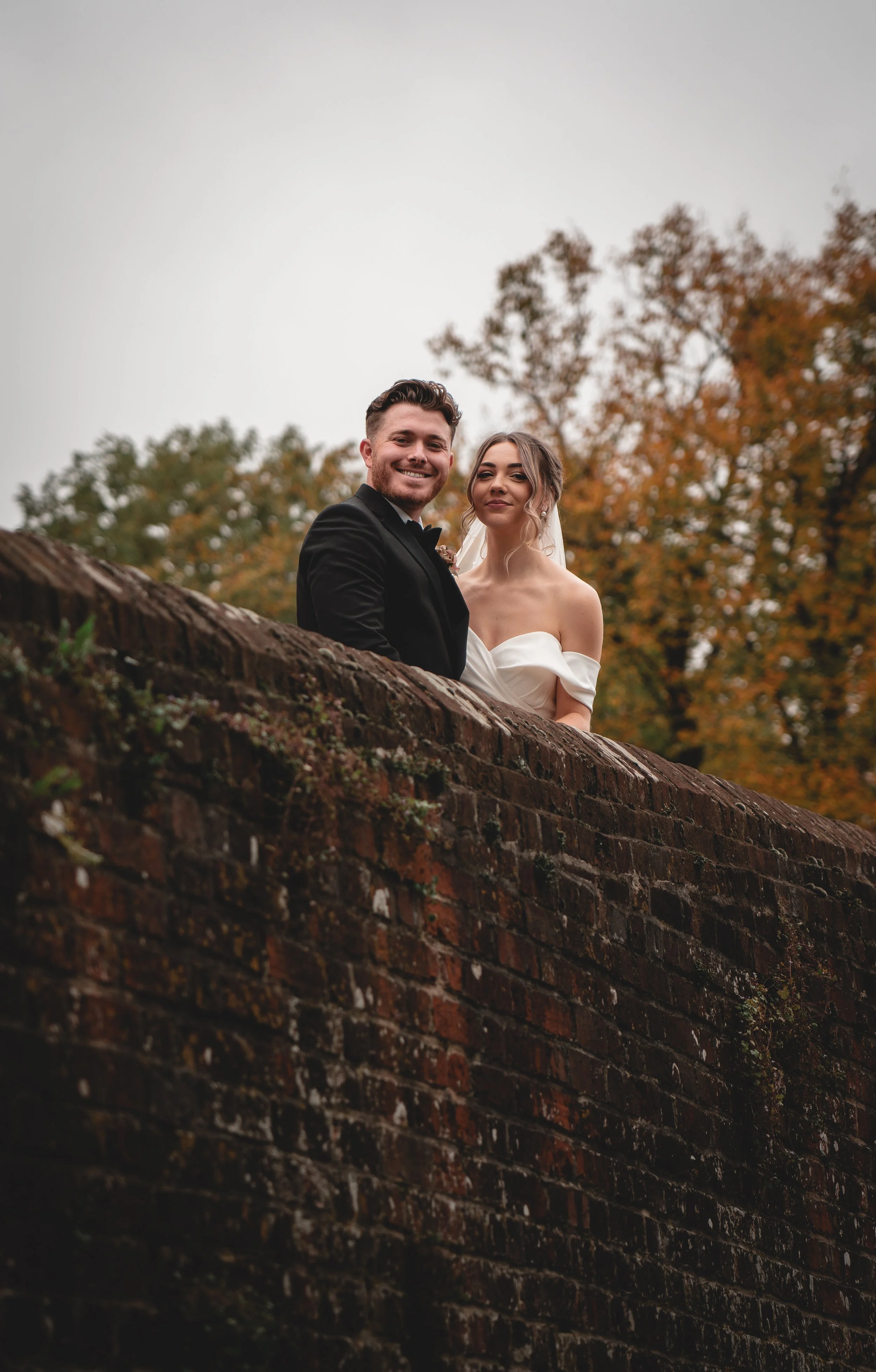A happy bride and groom on their wedding day standing behind a brick wall with autumn trees in the background.